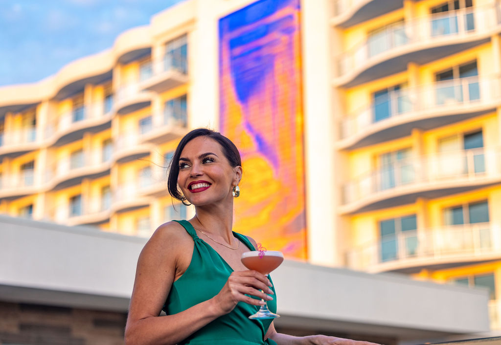 A woman in a green dress holds a cocktail and smiles, standing outdoors in front of a modern building with yellow balconies and a colorful vertical mural.