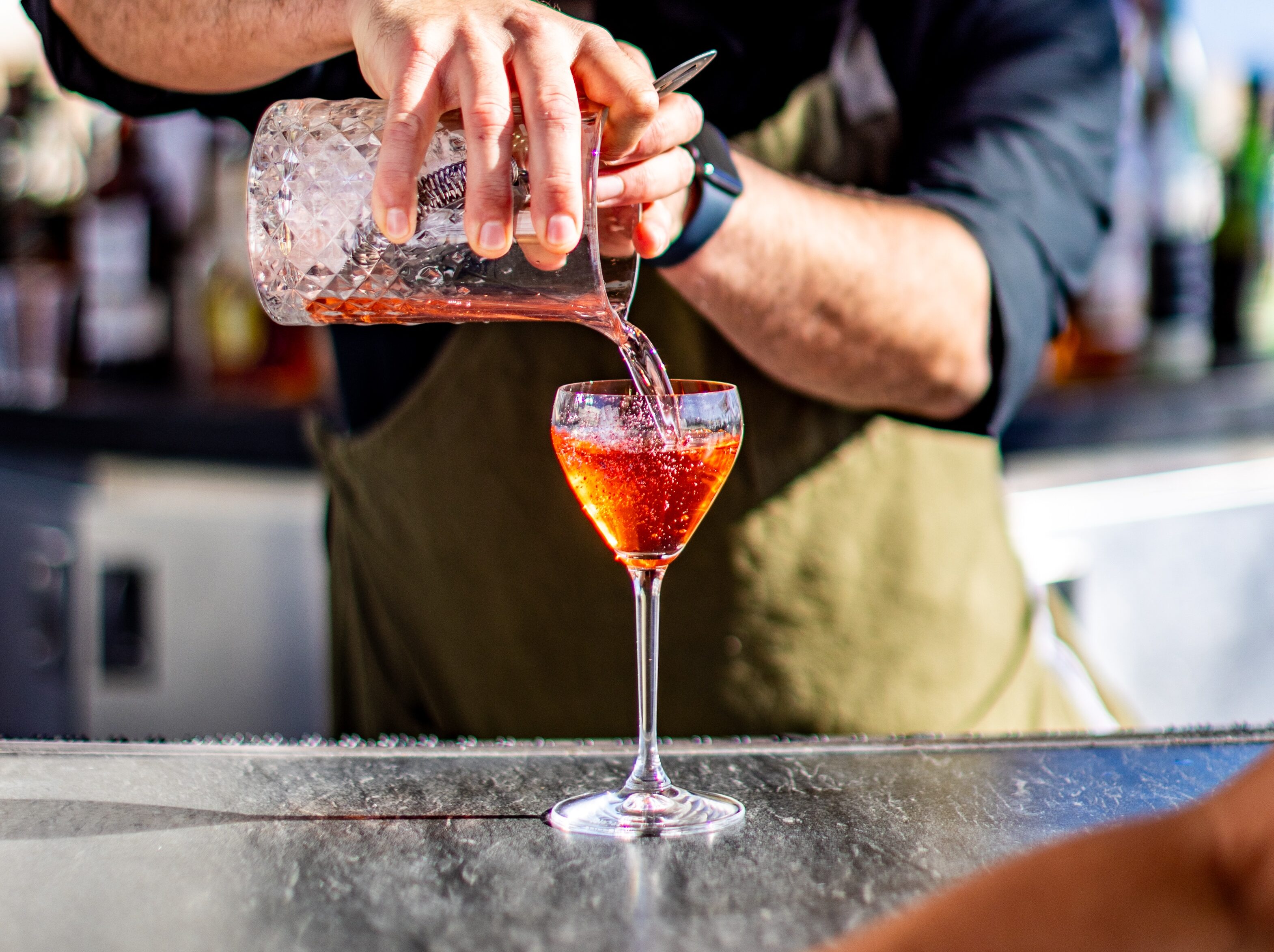 A bartender wearing sunglasses pours a red cocktail from a mixing glass into a stemmed glass at an outdoor bar.