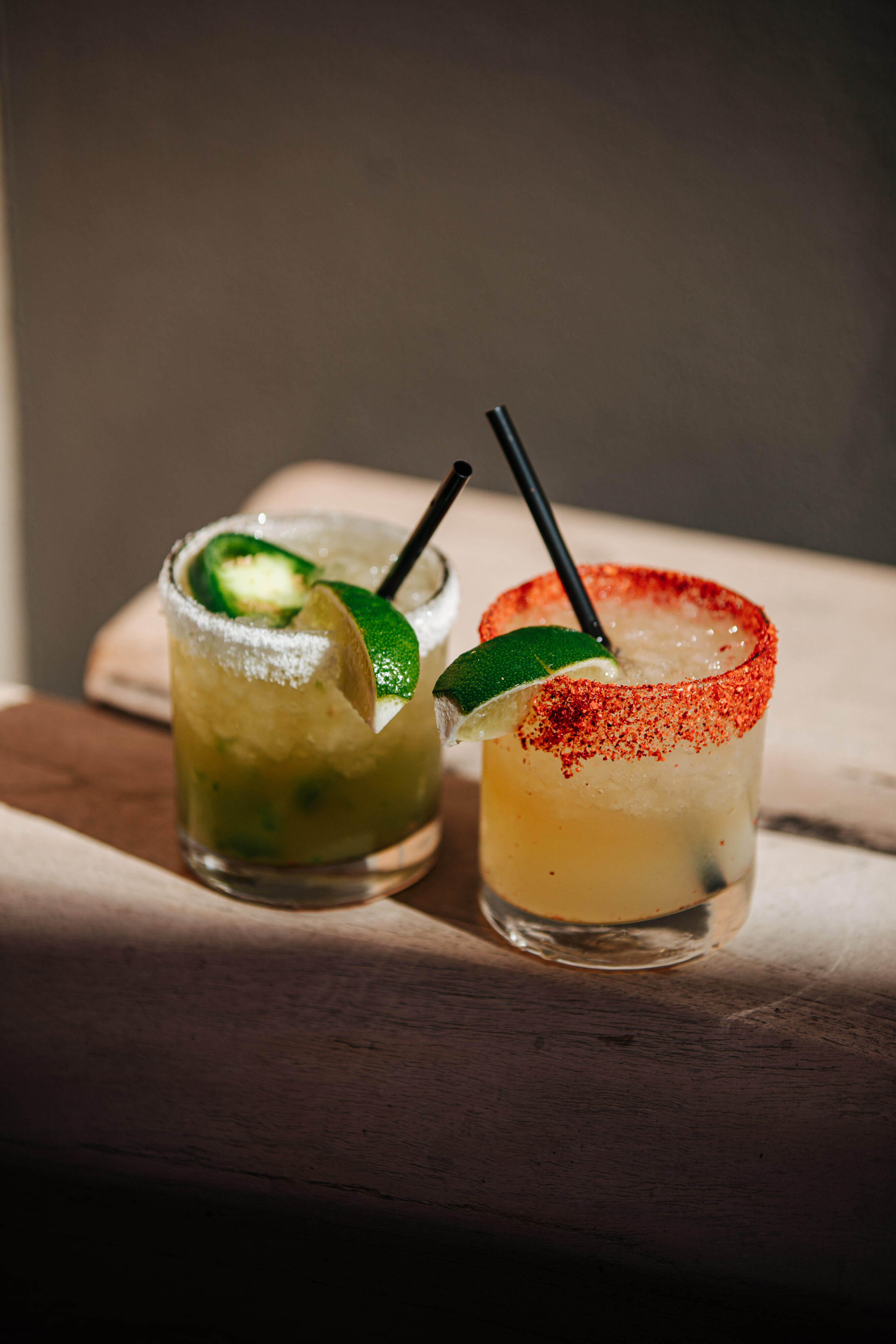 Two cocktails in short glasses with lime wedges, featured on our cocktail menu—one with a salted rim, the other dusted with red chili powder. Both have black straws and sit atop a light wooden surface.