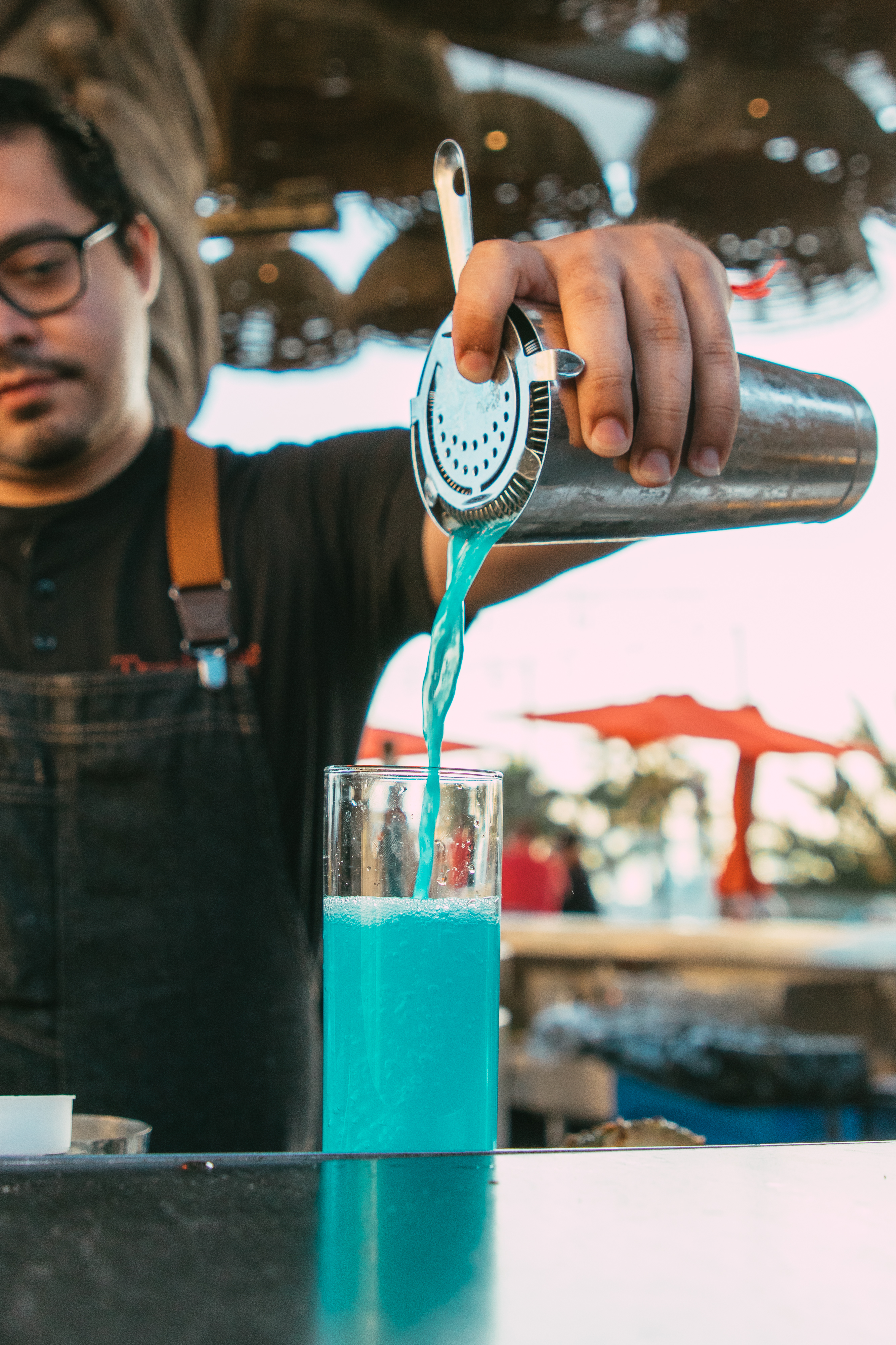 A bartender wearing glasses pours a bright blue cocktail from a shaker into a tall glass at an outdoor bar.
