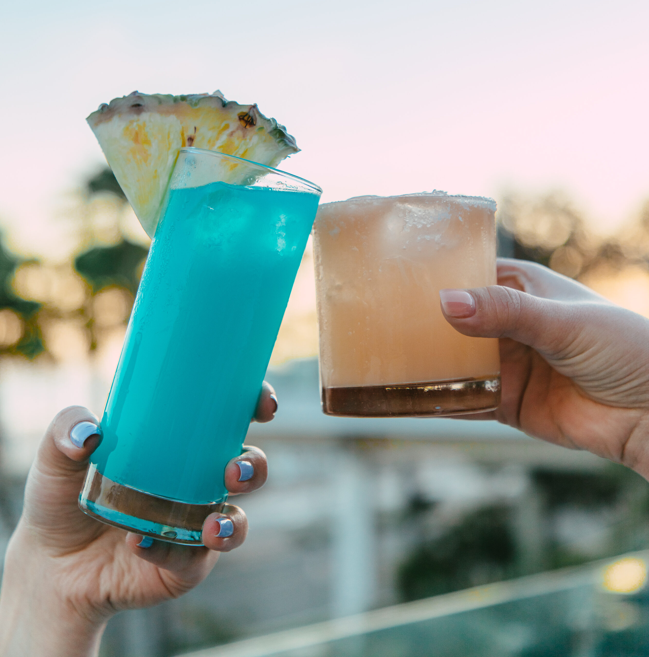 Two hands hold up different cocktails outdoors; one is blue with a pineapple slice, the other is light brown with a salted rim. Blurred background shows trees and sky.