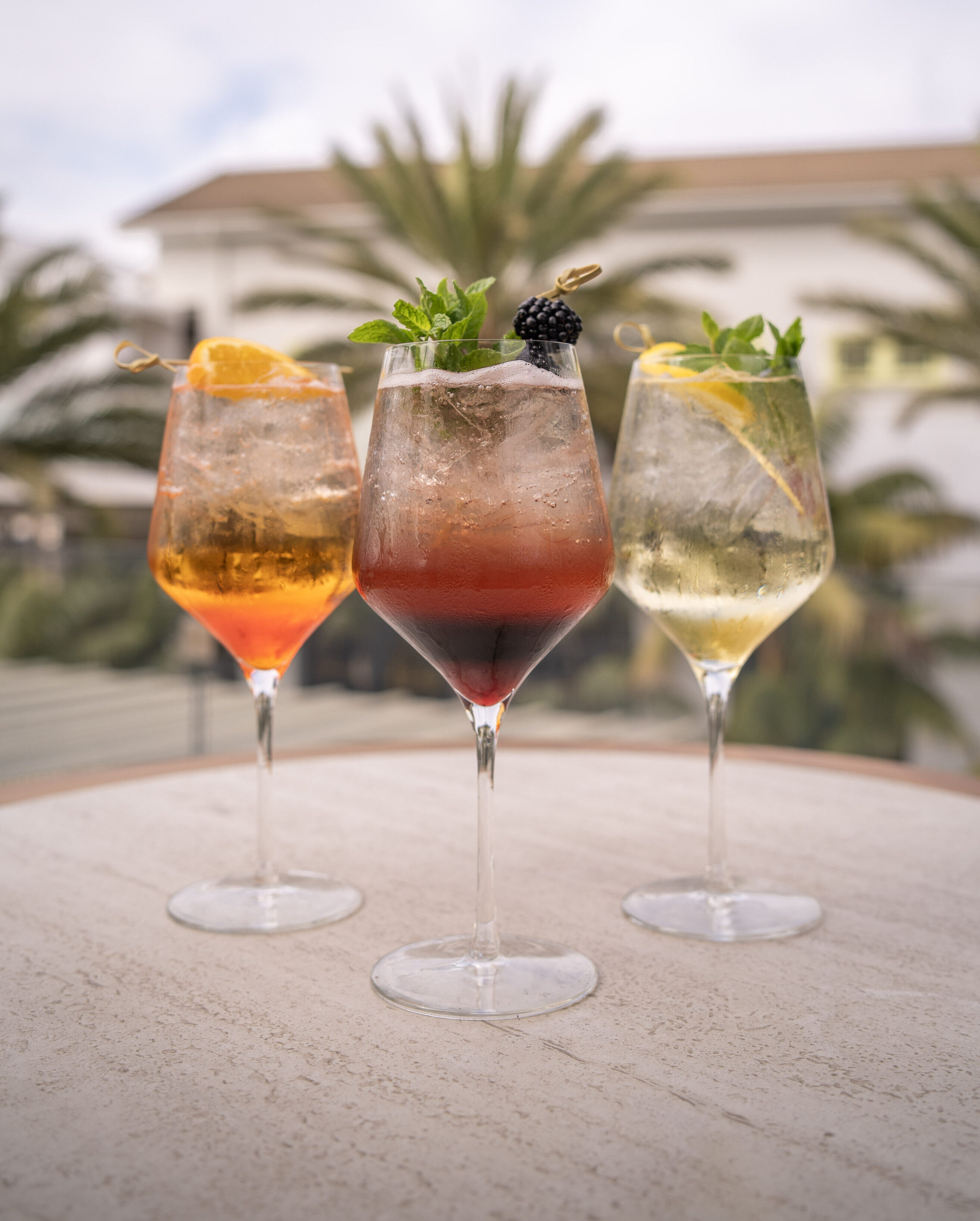 Three colorful cocktails in large wine glasses, garnished with fruit and herbs, are displayed on a round table outdoors with blurred palm trees in the background.
