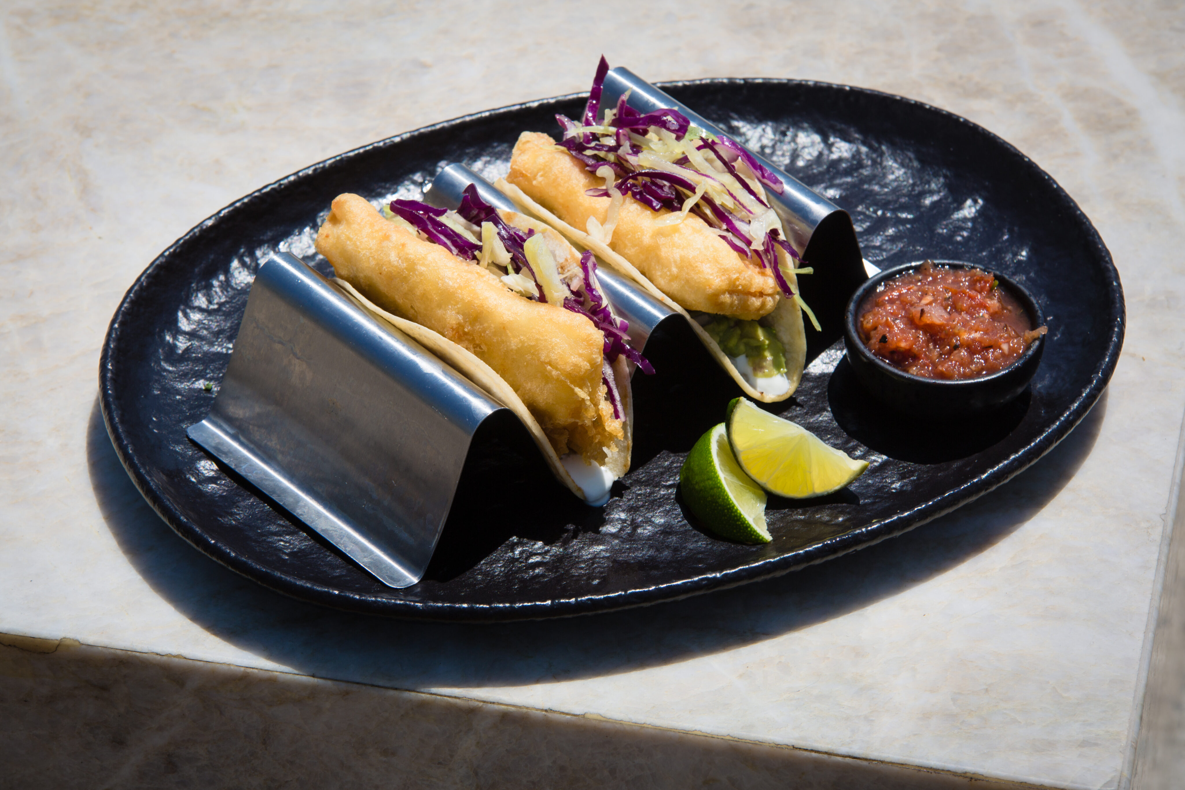 Two fish tacos with shredded cabbage in metal taco holders on a black plate, served with lime wedges and a small cup of salsa.