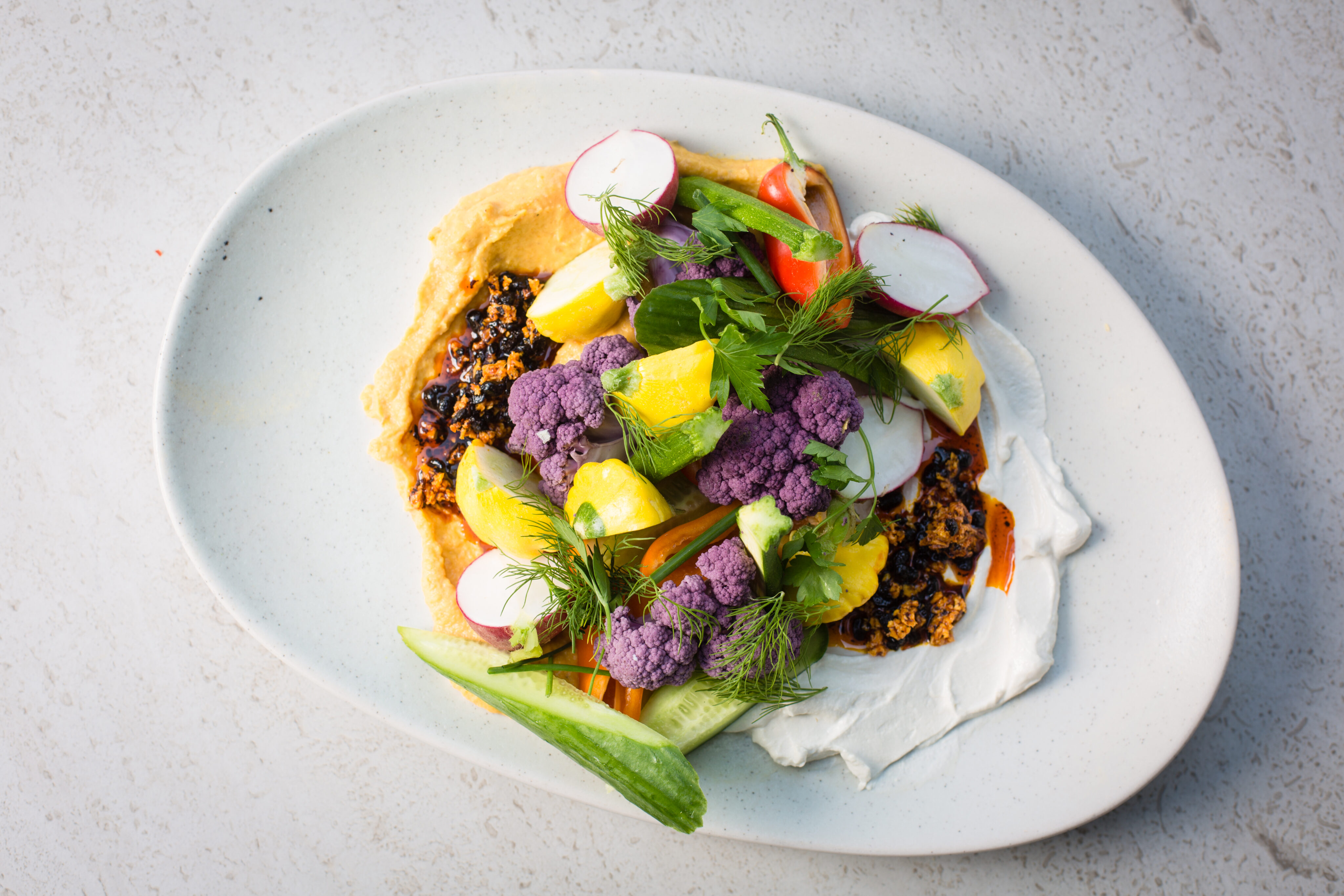 A white oval plate with hummus, white spread, black quinoa, purple cauliflower, pineapple, cucumber, radish, carrots, dill, and greens arranged on top.