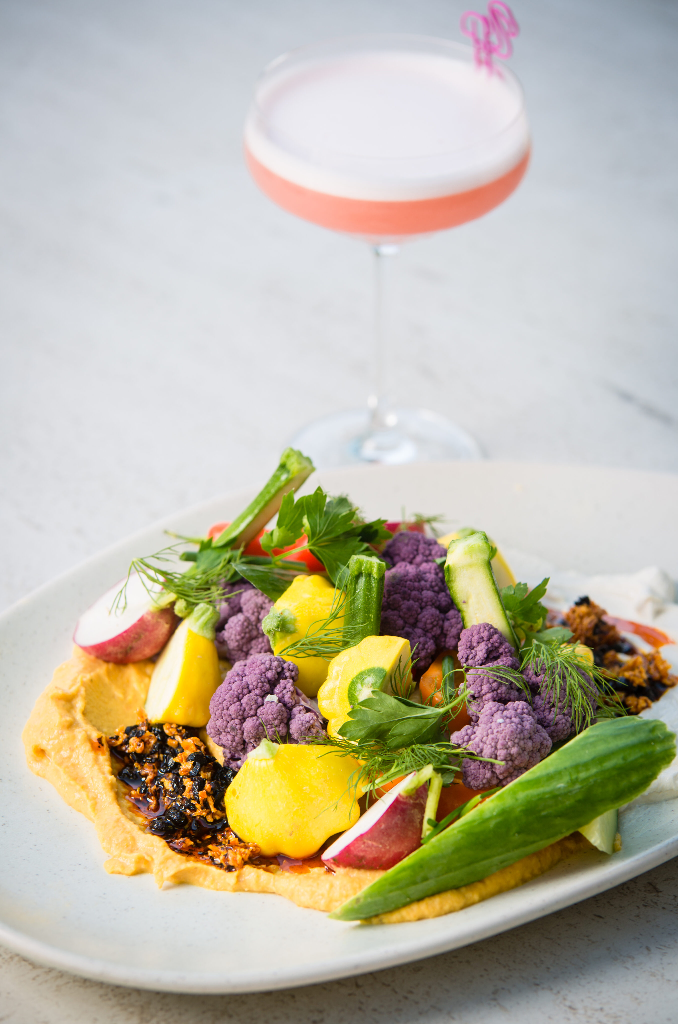 A plate of assorted fresh vegetables on a creamy spread, garnished with herbs, with a pink cocktail in a glass in the background.