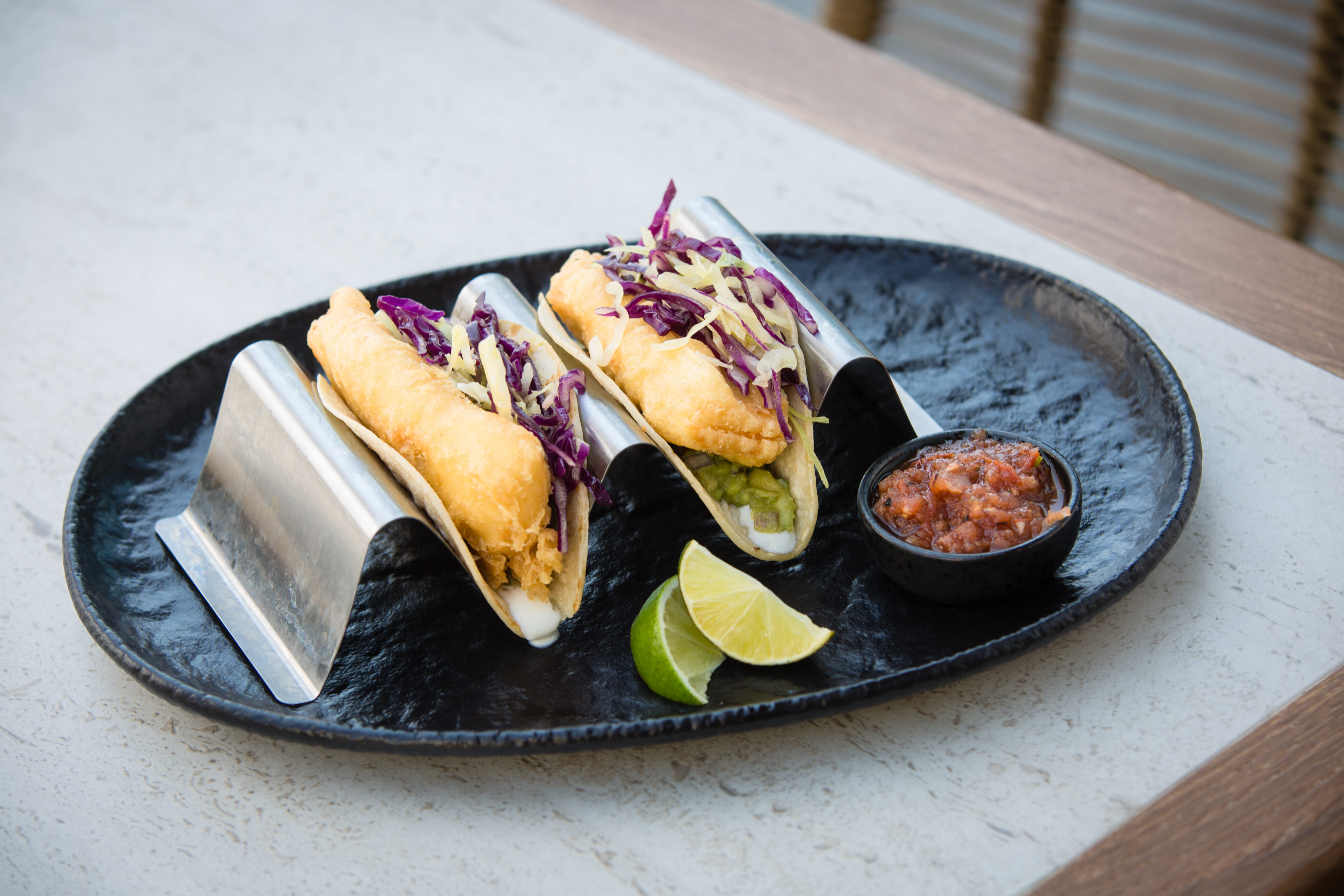 Two battered fish tacos with cabbage and cheese on metal taco holders, served with lime wedges and salsa on a black oval plate.