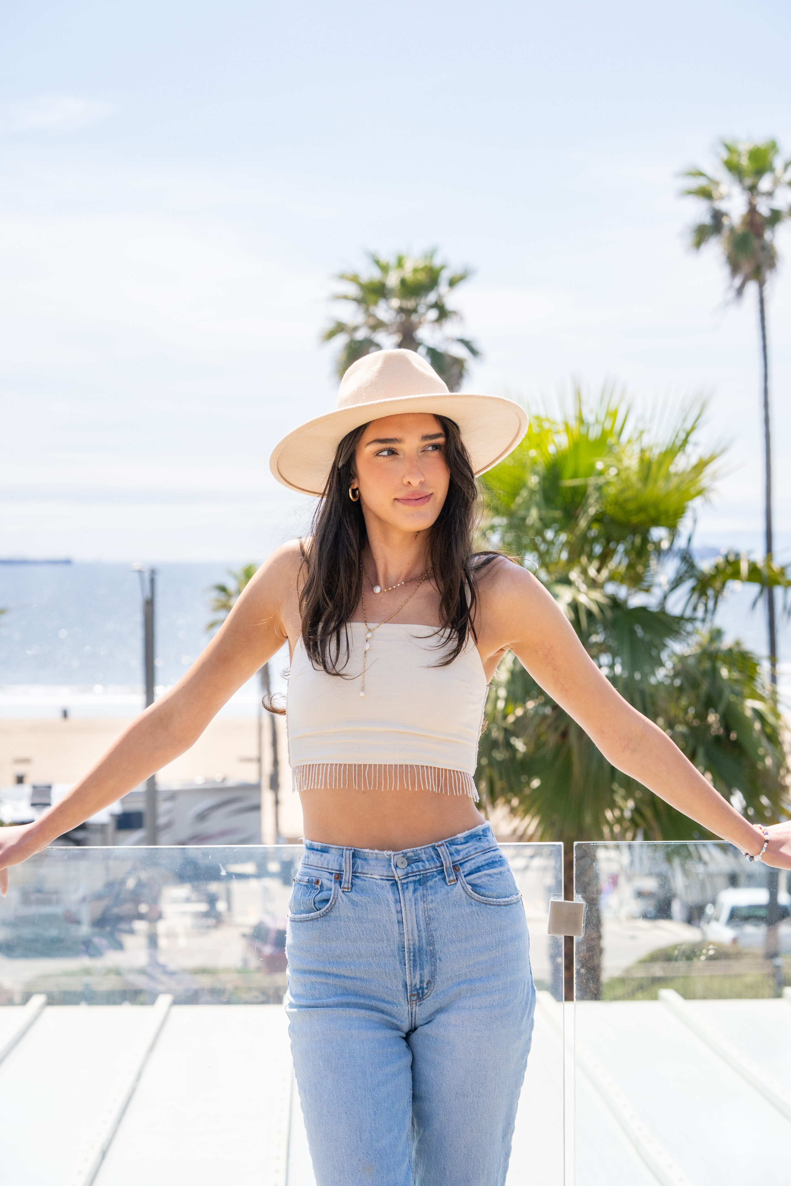 A woman wearing a wide-brimmed hat, a beige crop top, and jeans stands outdoors by a glass railing with palm trees and the ocean in the background.