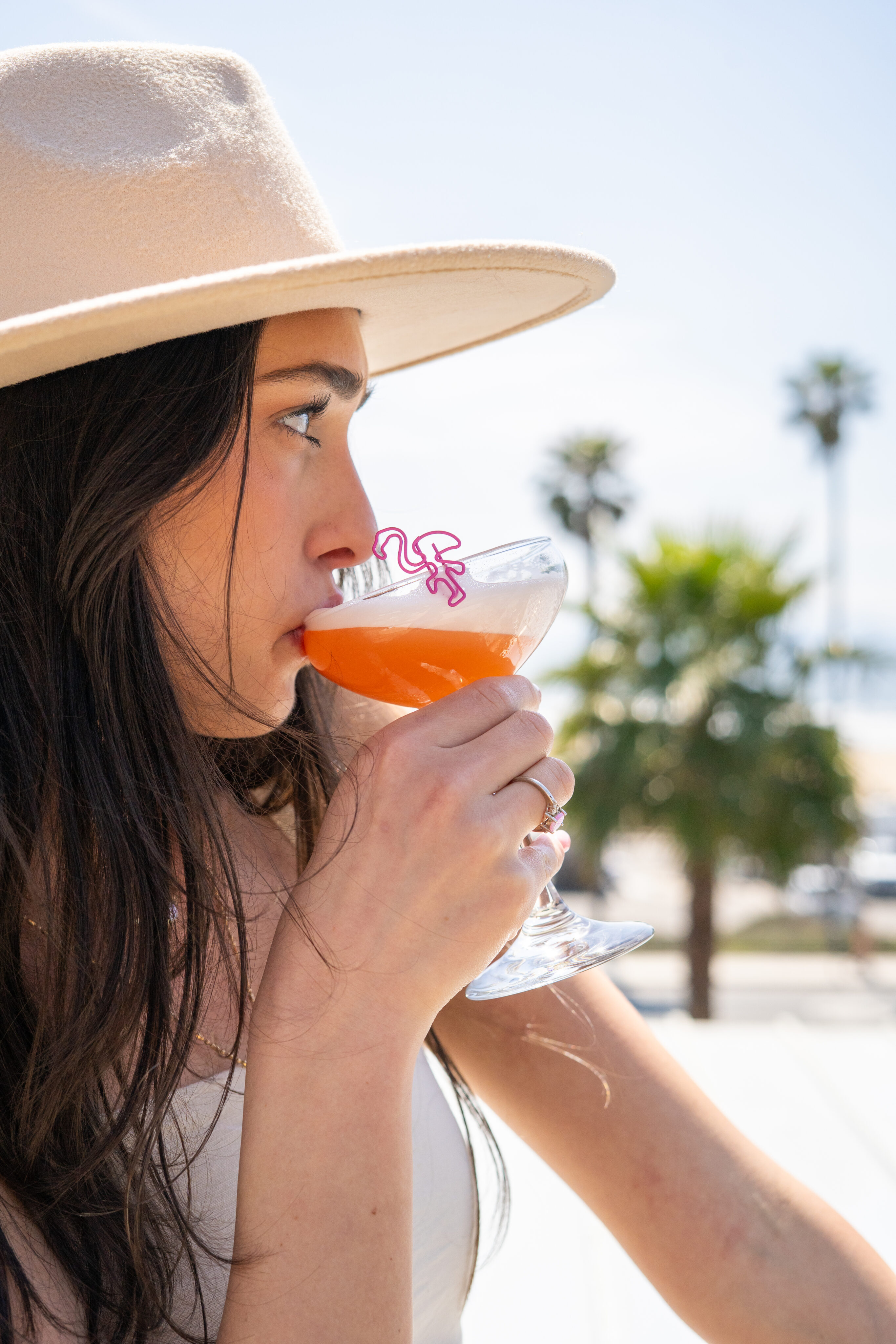 A woman in a beige hat enjoys an orange cocktail made with Código 1530 Tequila and a pink flamingo straw, with palm trees and a bright sky setting the scene for a perfect Blanco Party.