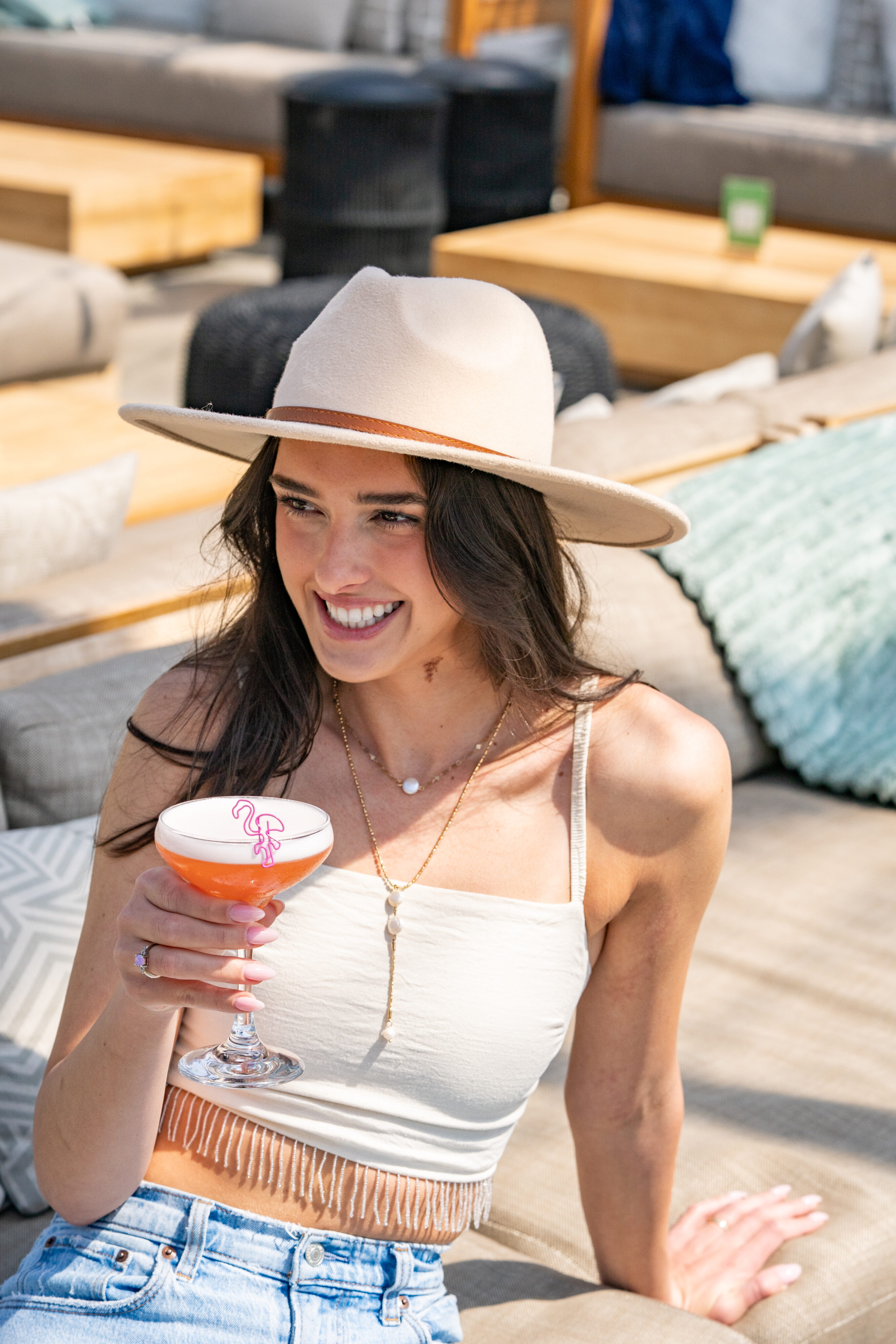 A woman in a hat and white top sits on an outdoor sofa, smiling as she enjoys a pink cocktail with foam art—perfect for a sunny Weekend Brunch surrounded by stylish lounge furniture.