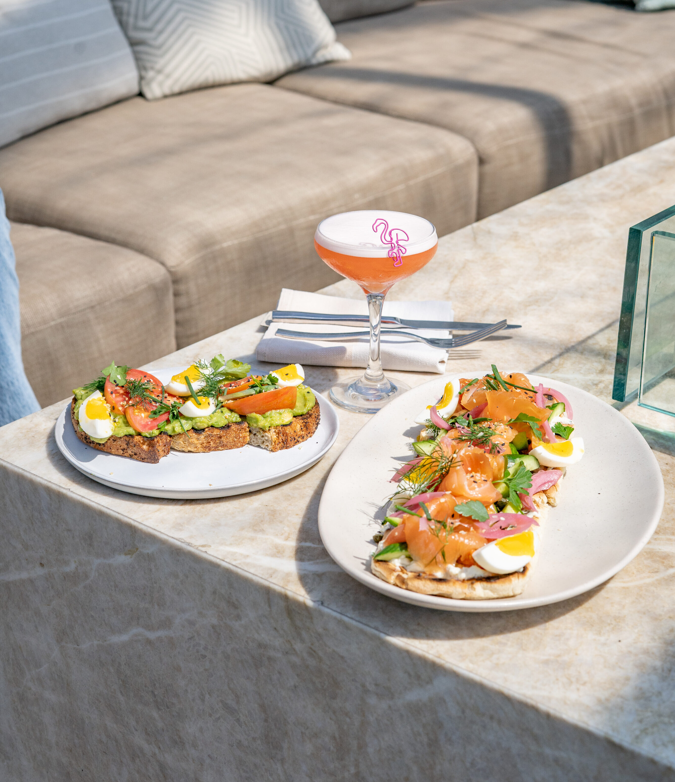 Two plates of open-faced sandwiches with eggs and smoked salmon, plus a pink cocktail, invite you to relax outdoors on cushioned lounge seating—the perfect setting for a leisurely weekend brunch.