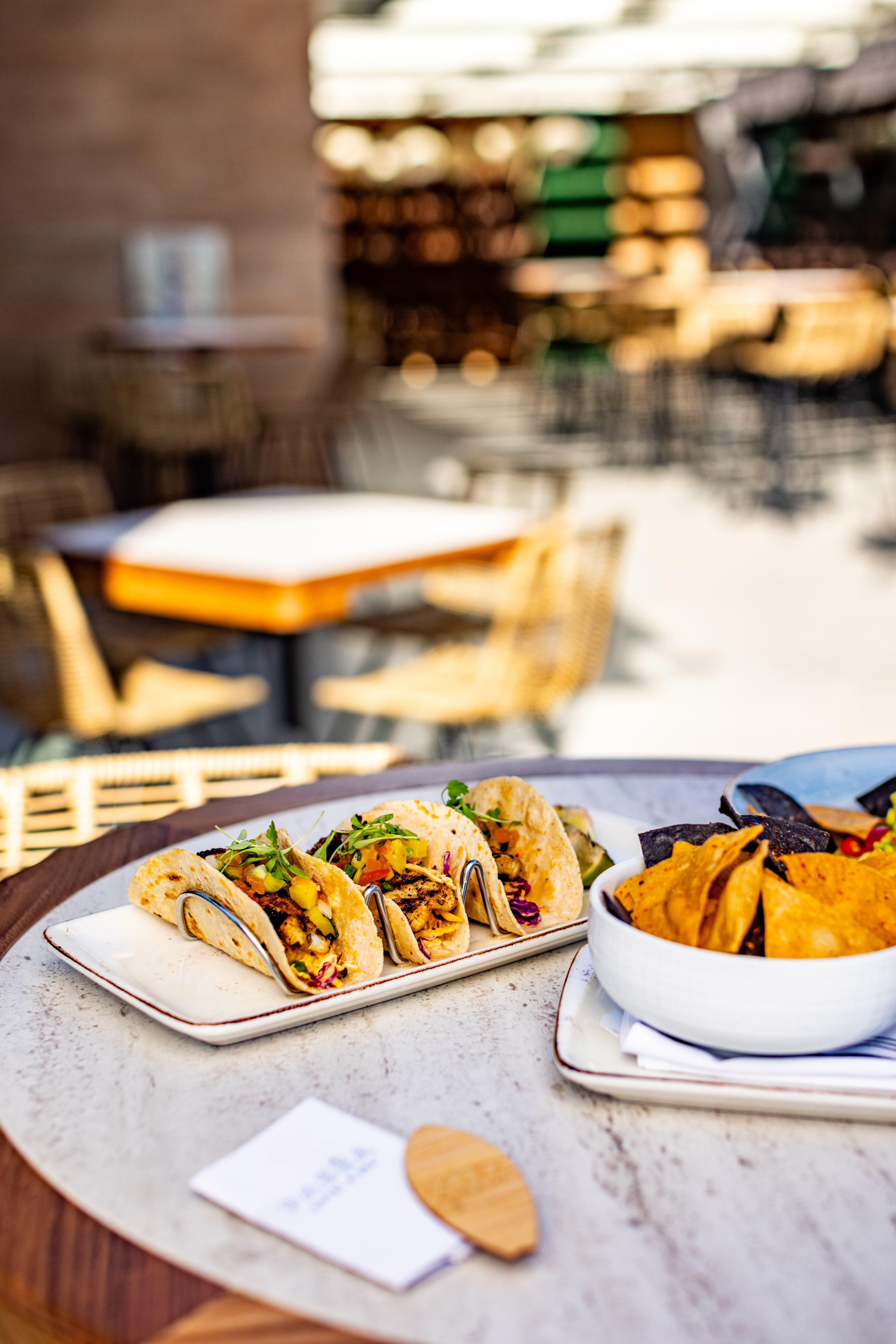 Three tacos with various fillings are served on a rectangular plate next to a bowl of tortilla chips on an outdoor restaurant table.