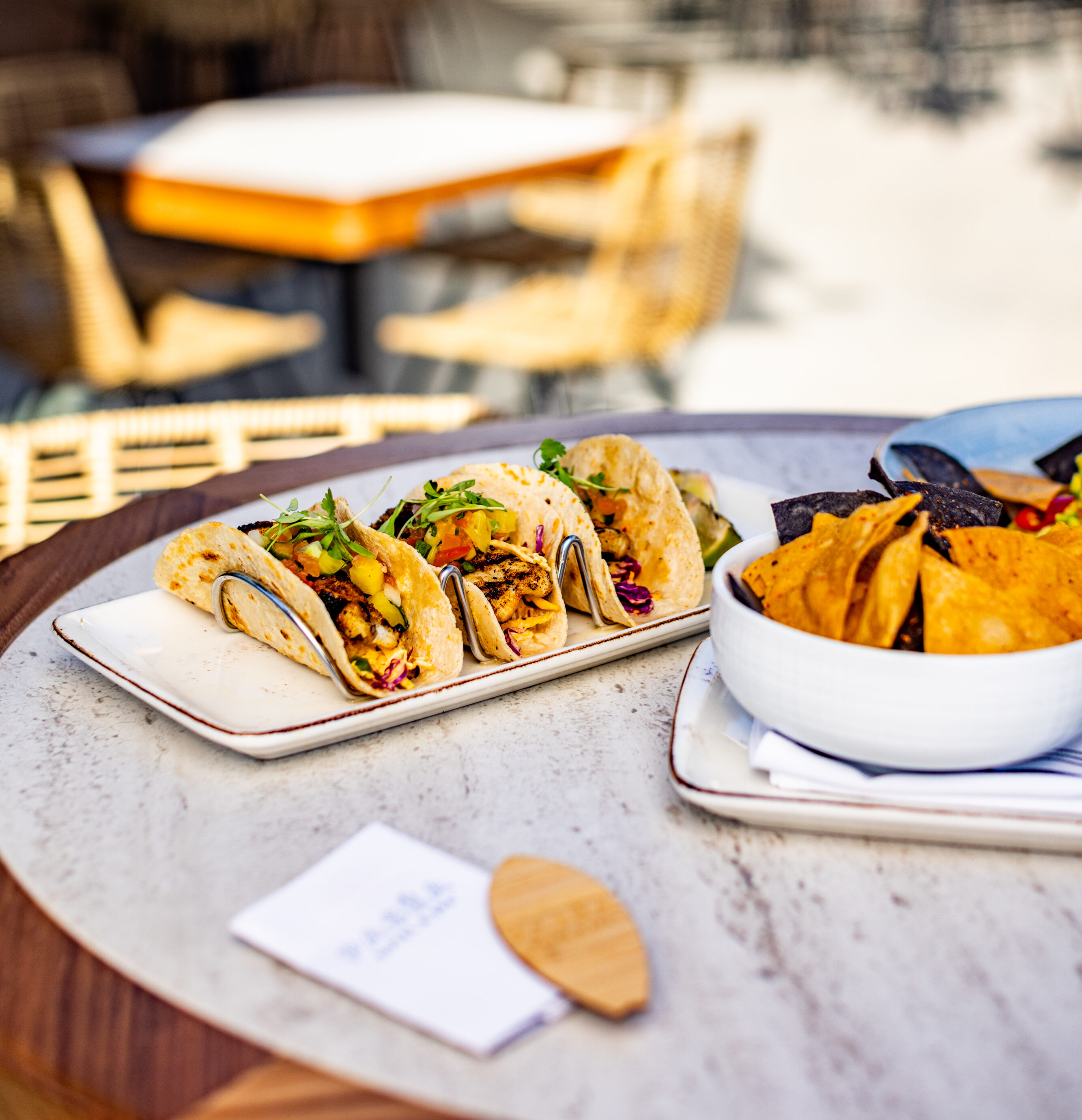 A plate with three tacos and a bowl of tortilla chips are set on a round table at an outdoor dining area with empty chairs and tables in the background.