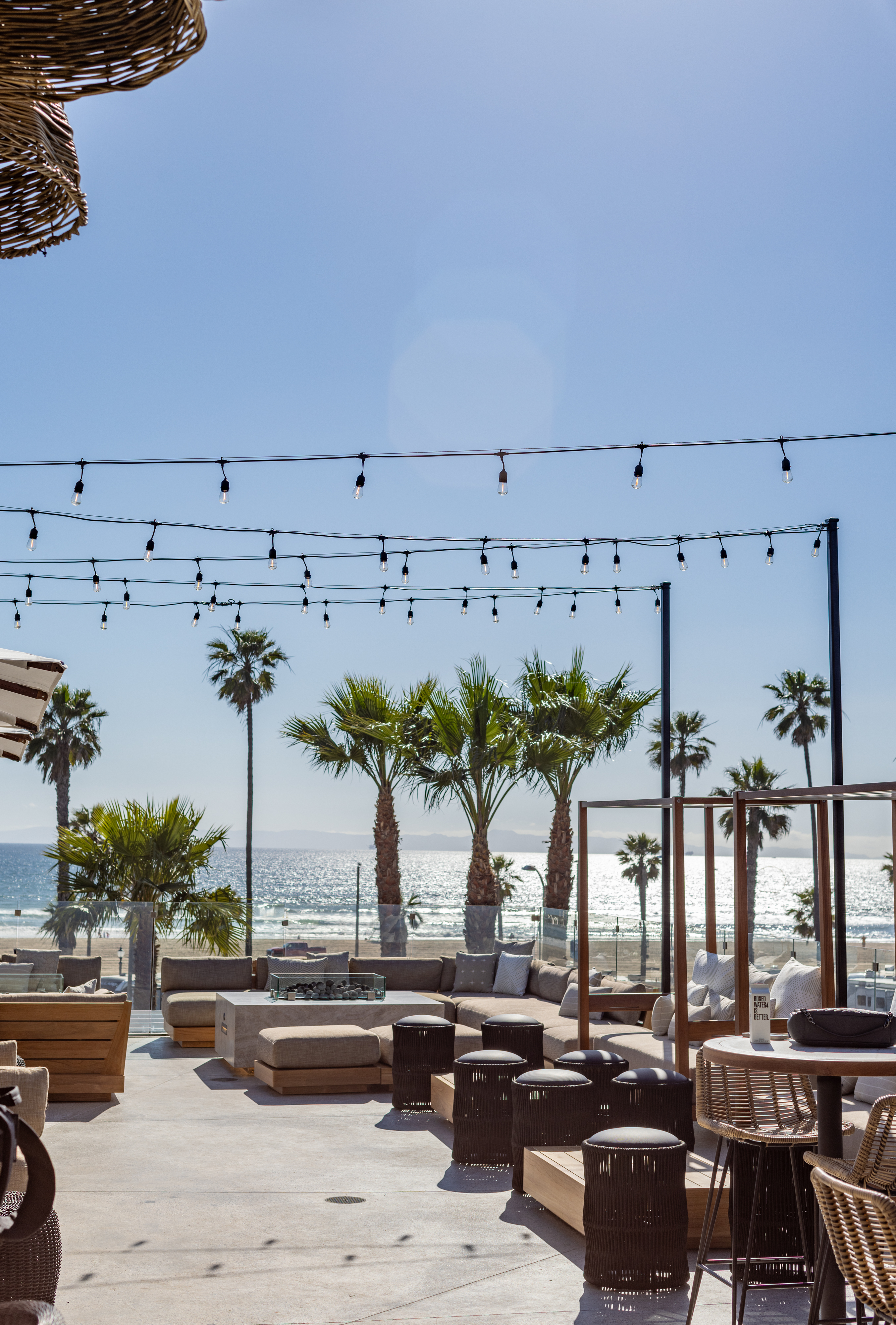 Outdoor seating area with wooden furniture, string lights overhead, palm trees, and an ocean view on a sunny day.