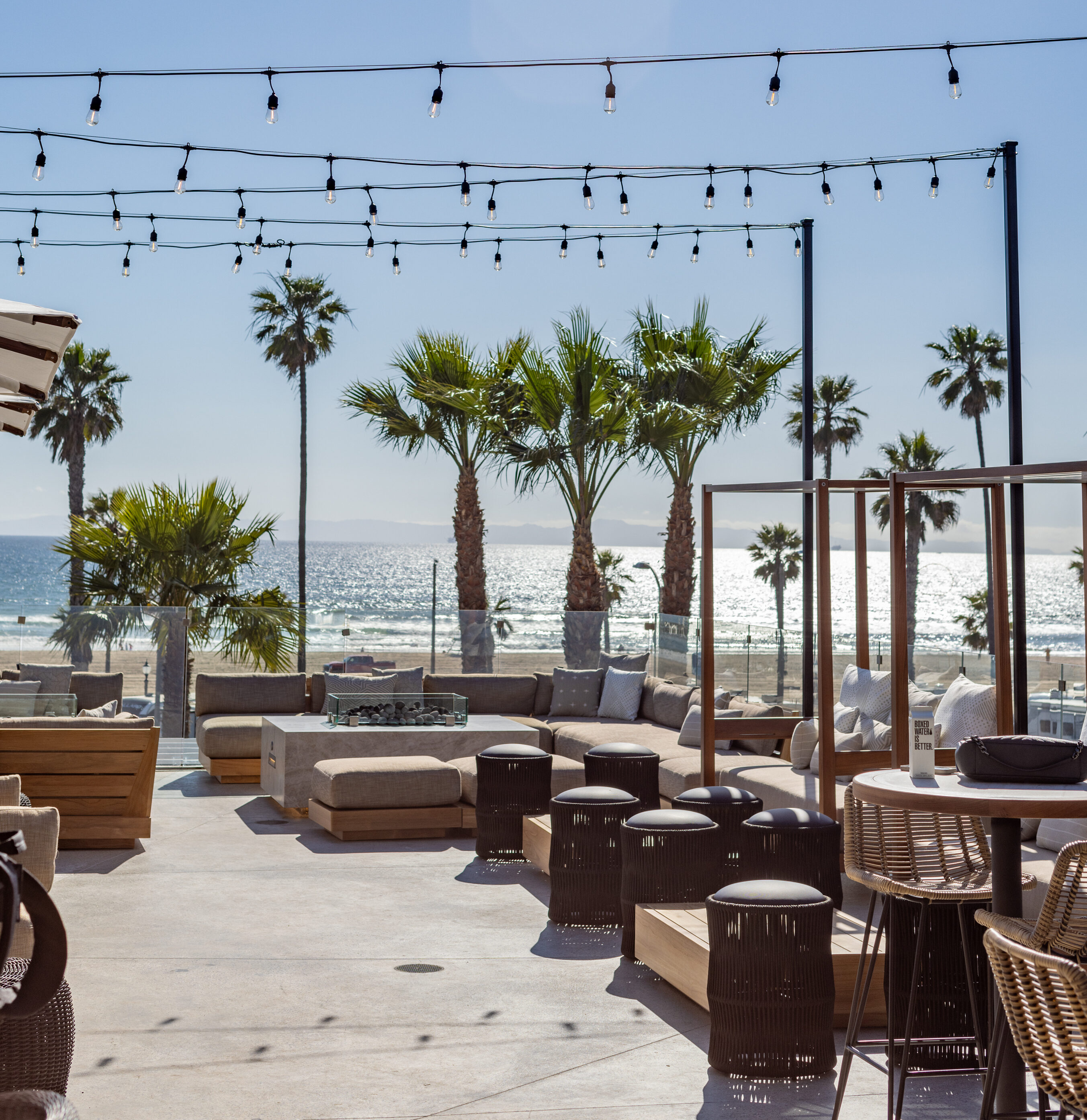 Outdoor beachside patio with lounge seating, string lights, palm trees, and a view of the ocean under a clear blue sky.