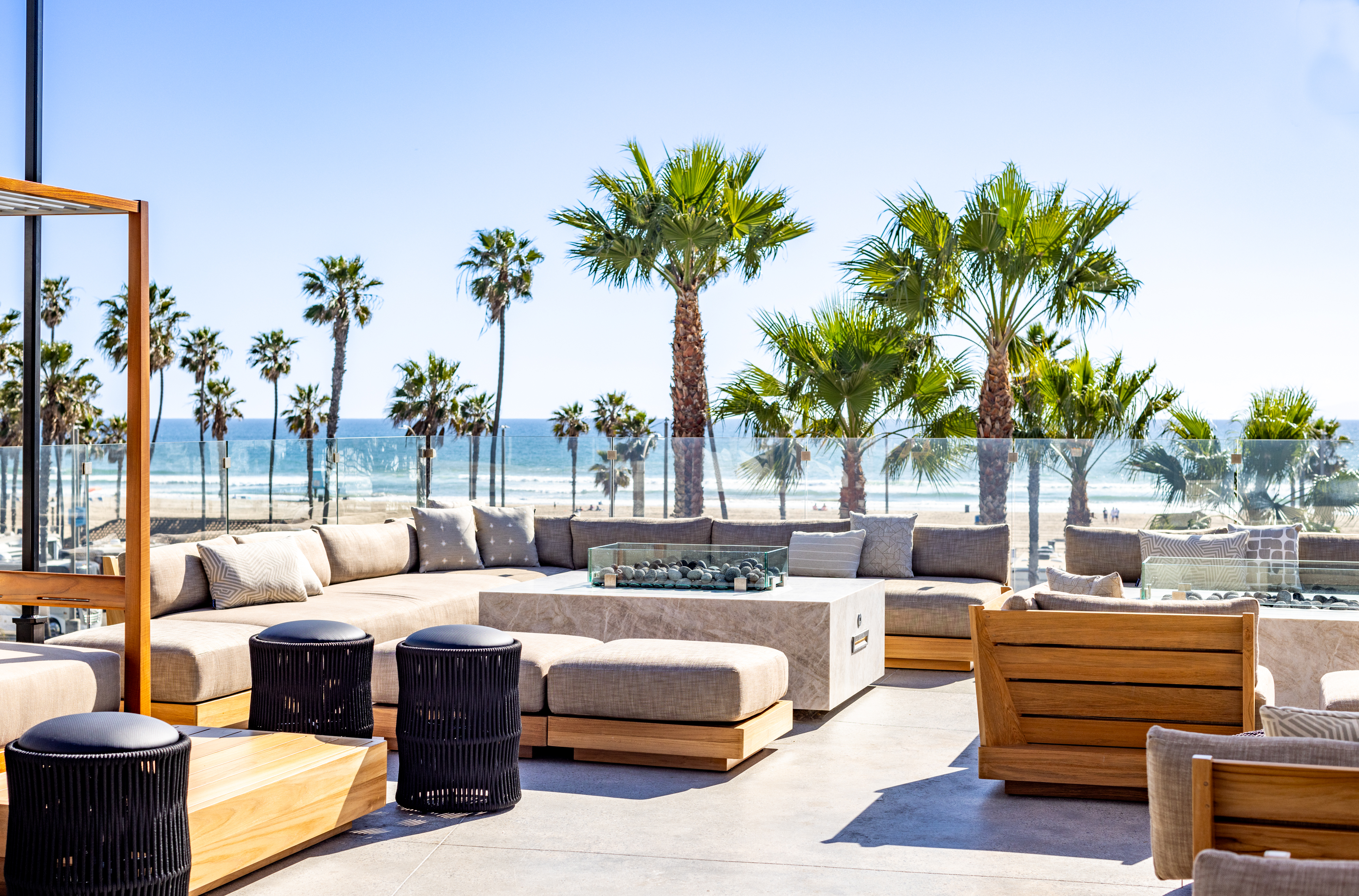 Outdoor lounge area with cushioned couch seating, wooden furniture, and fire pit, set beneath a cabana and overlooking a beach lined with palm trees under a clear blue sky.