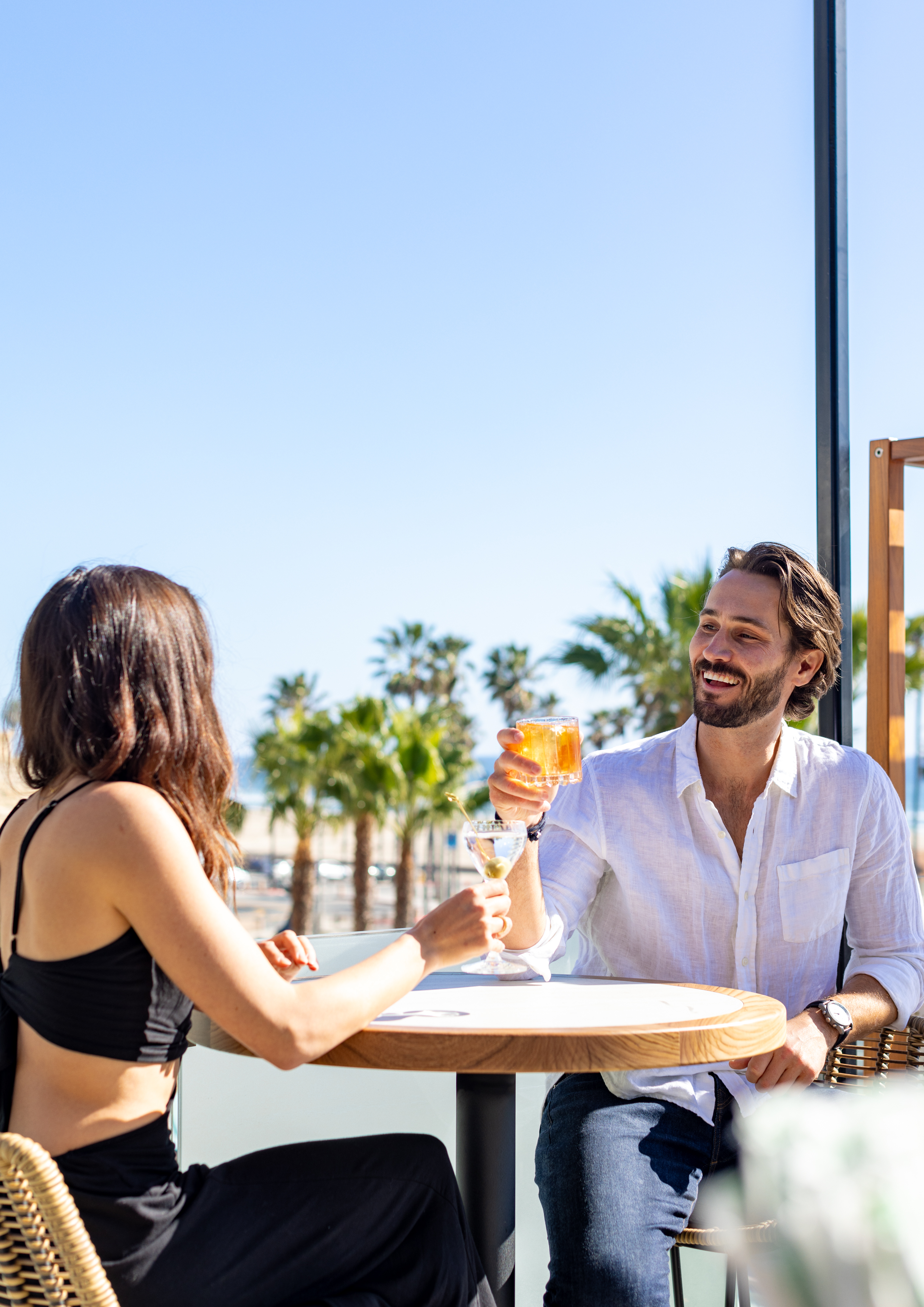 A man and woman sit at an outdoor table, clinking glasses and smiling, with palm trees and a clear blue sky in the background.