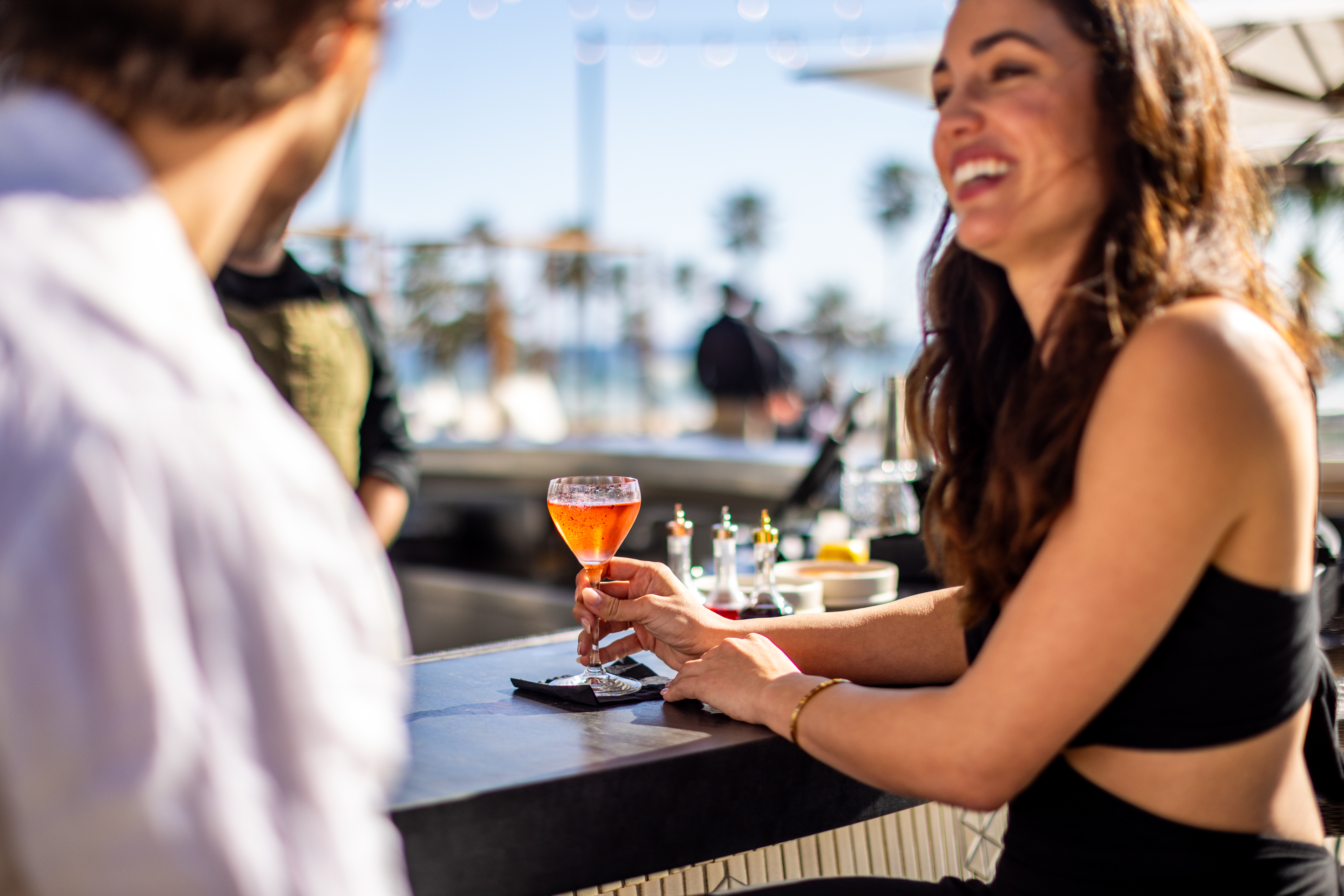 A woman holding a cocktail sits at an outdoor bar, smiling and talking with a man; palm trees and people are visible in the sunny background.