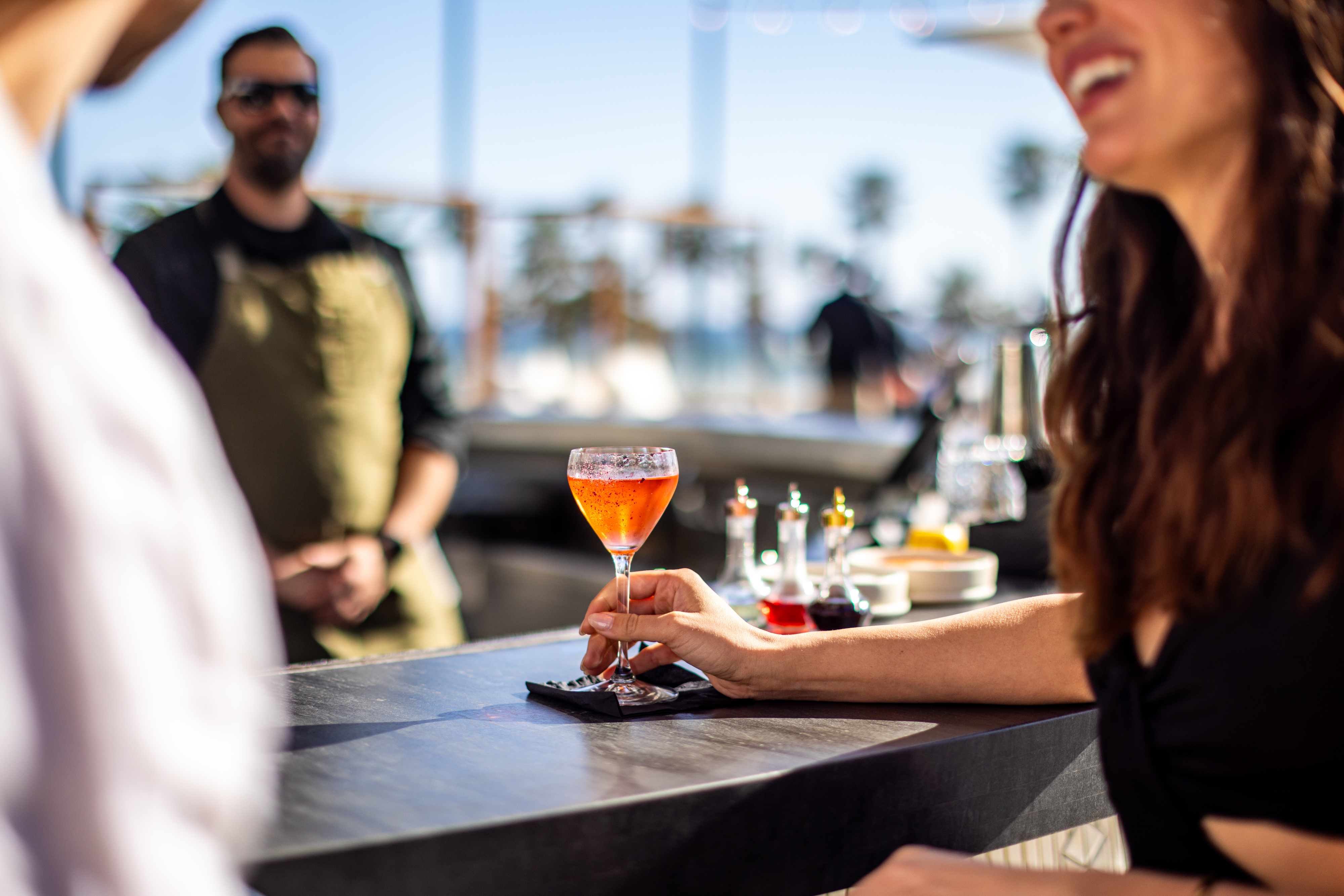A person holds a cocktail at an outdoor bar counter, with another person standing nearby and blurred background elements, including bottles and palm trees.