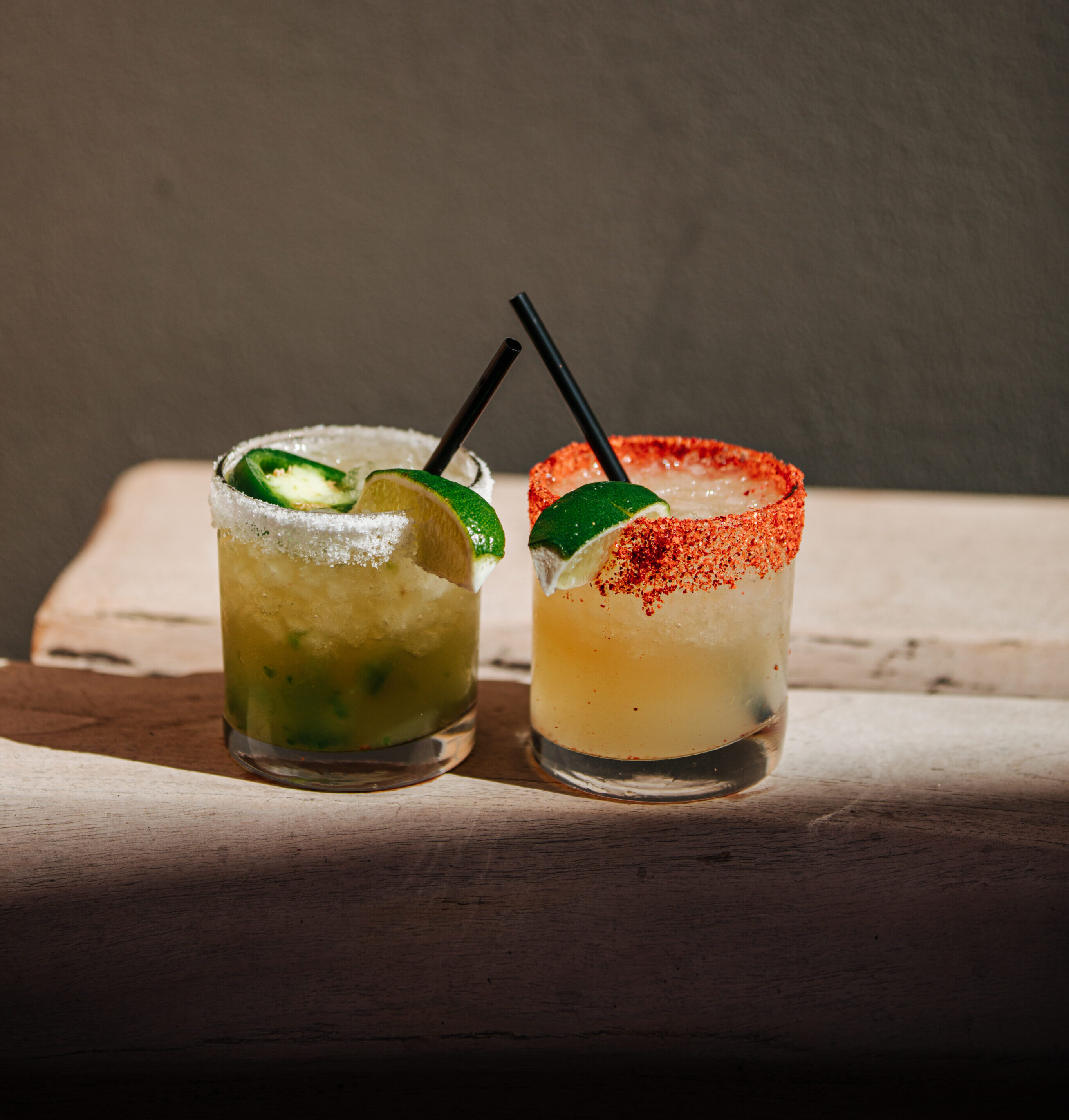 Two cocktails in short glasses on a light surface; one with jalapeño and salt rim, the other with chili powder rim and a lime wedge.