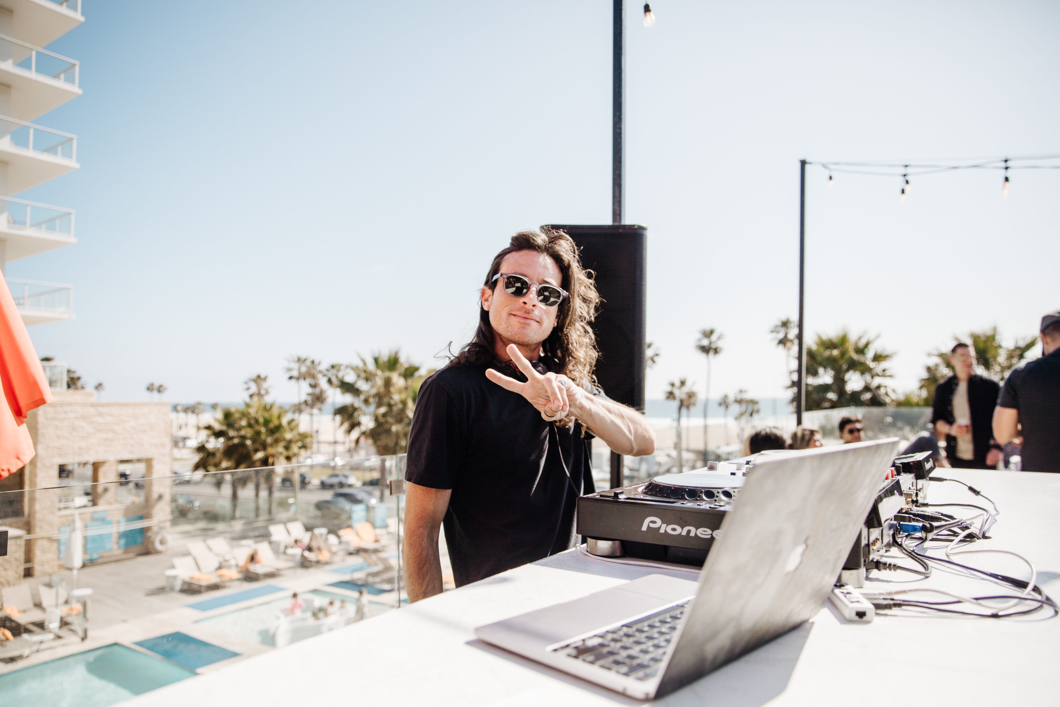 A DJ wearing sunglasses stands at an outdoor booth, making a peace sign near audio equipment with a laptop. A pool and palm trees are visible in the background.