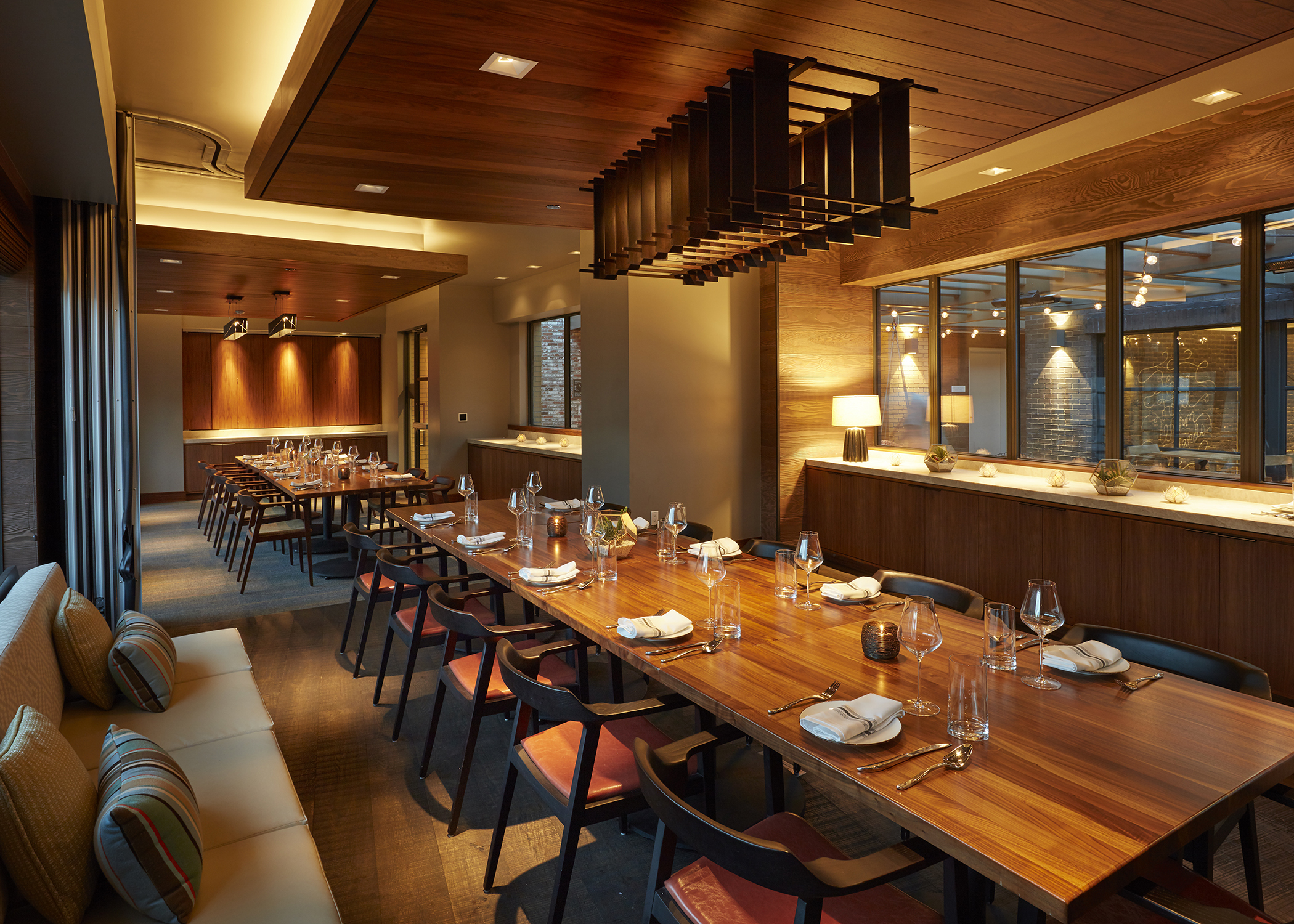 Modern dining room with long wooden tables, neatly arranged place settings, cushioned bench seating, and large windows letting in natural light. Warm ambient lighting and coastal touches create a cozy atmosphere perfect for drifting into relaxation.