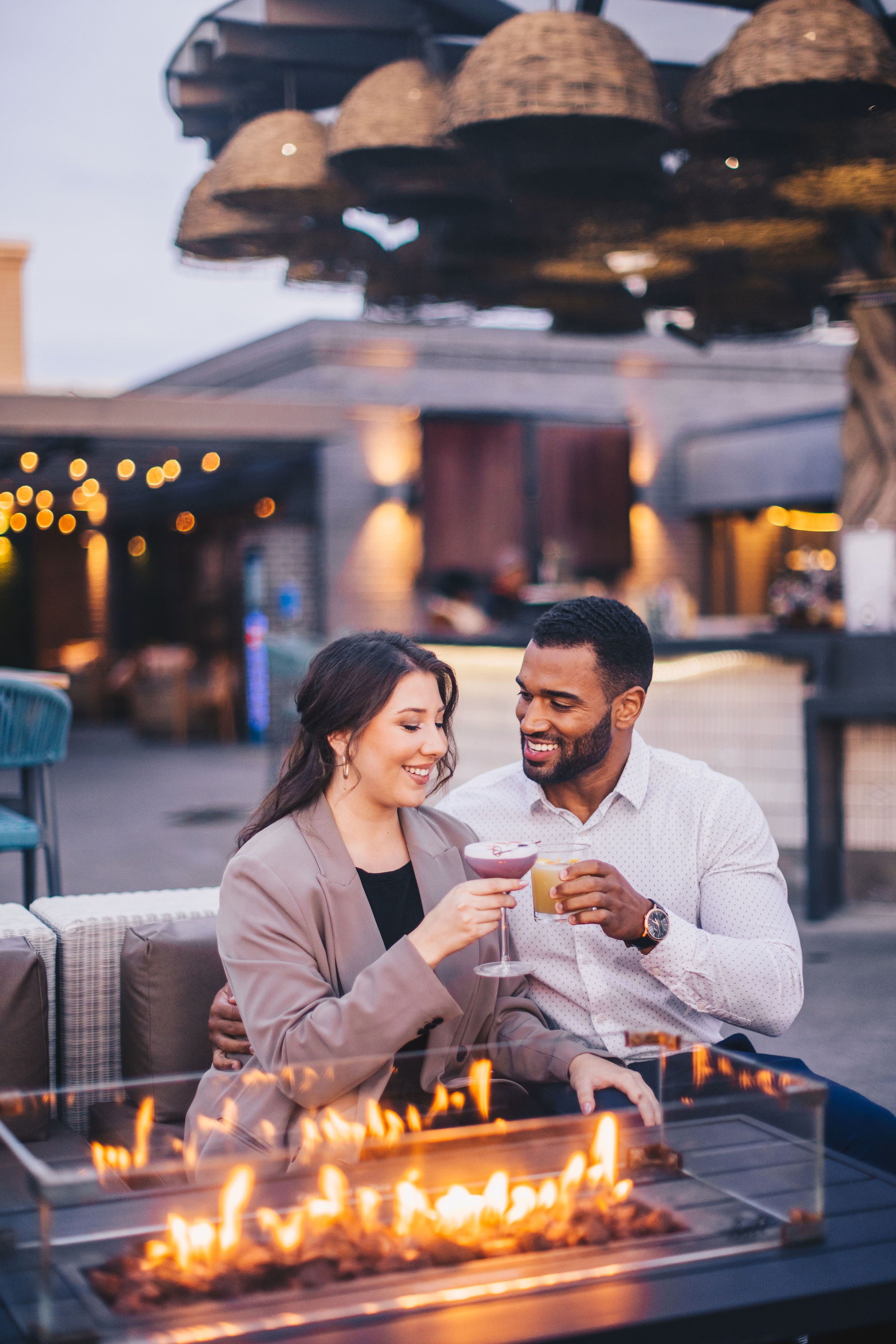 A man and woman sit together by a fire pit at an outdoor lounge, smiling and holding drinks, with warm lighting and patio decor in the background.