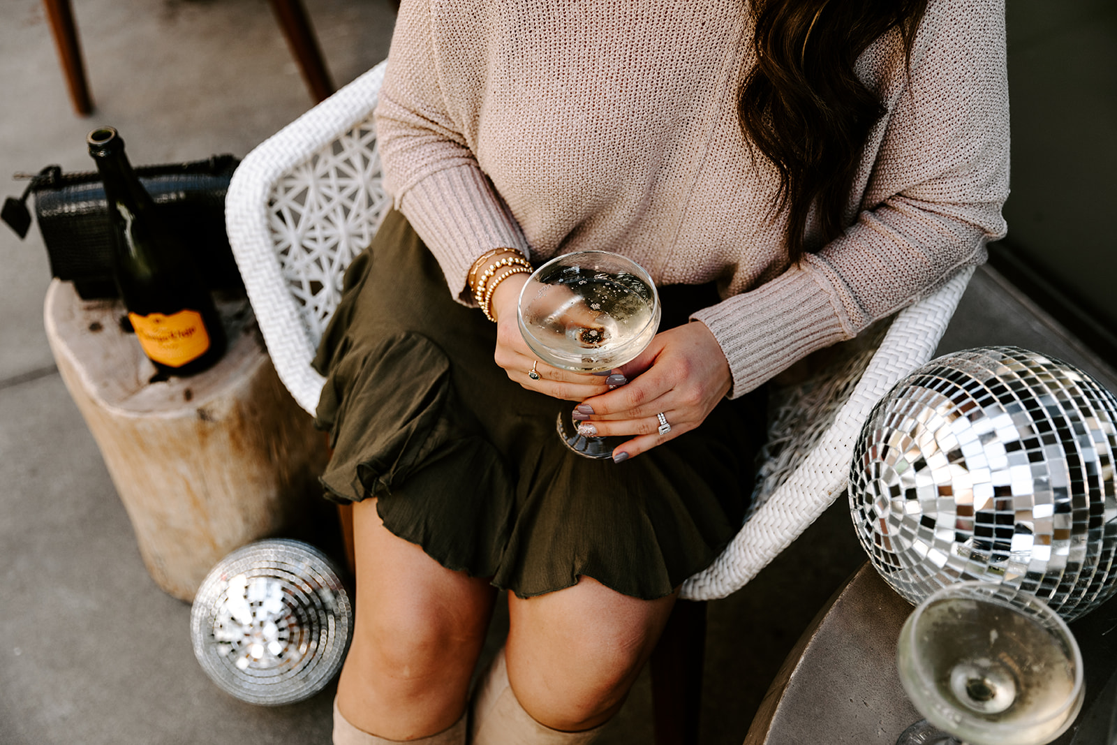 A person sitting on a white chair holds a glass of sparkling wine, surrounded by a bottle, disco balls, and a purse—capturing the festive vibe of a New Year's Eve Party.