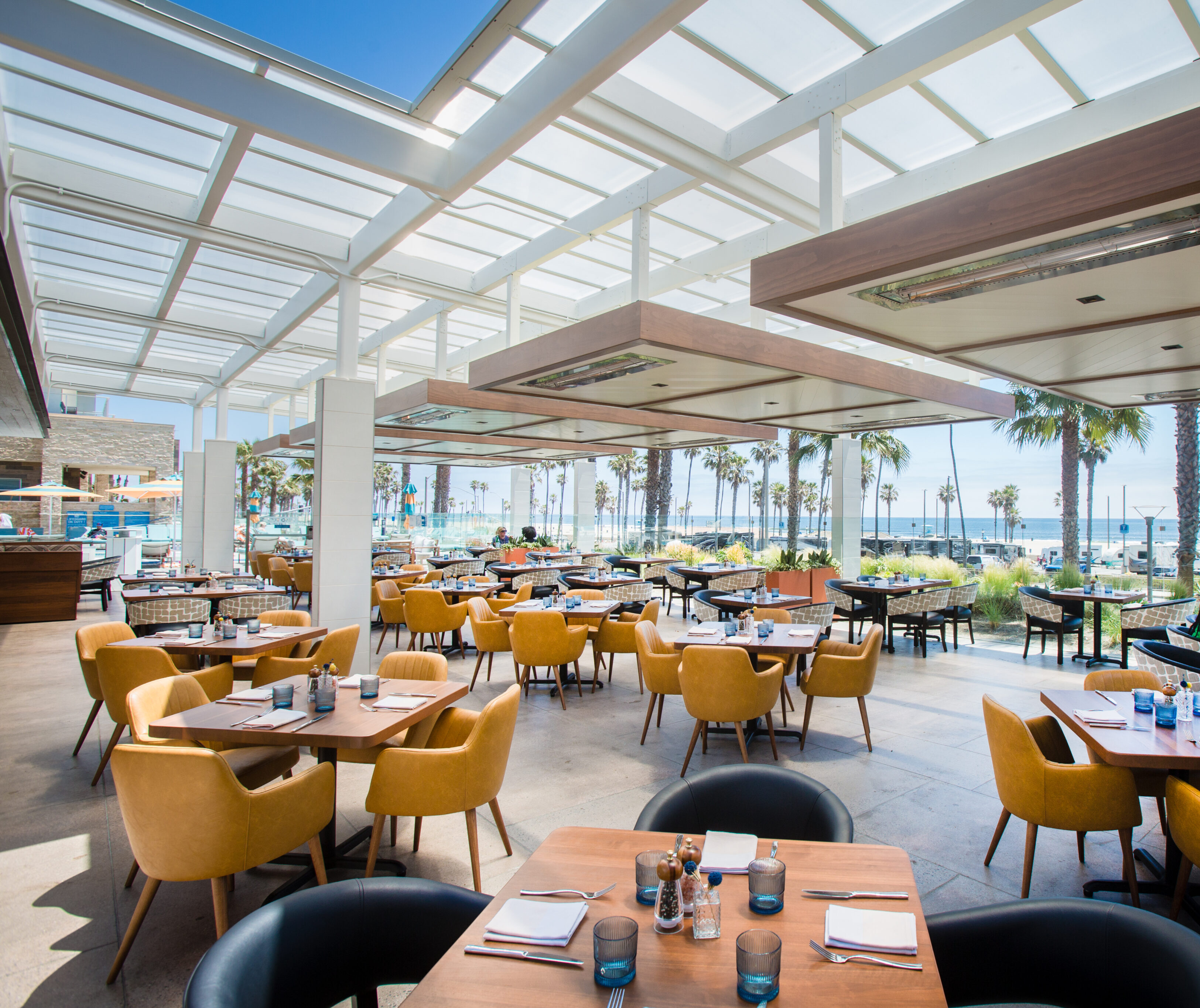 Modern outdoor restaurant with yellow and black chairs, set tables, and a view of palm trees and the ocean through glass walls under a white pergola.