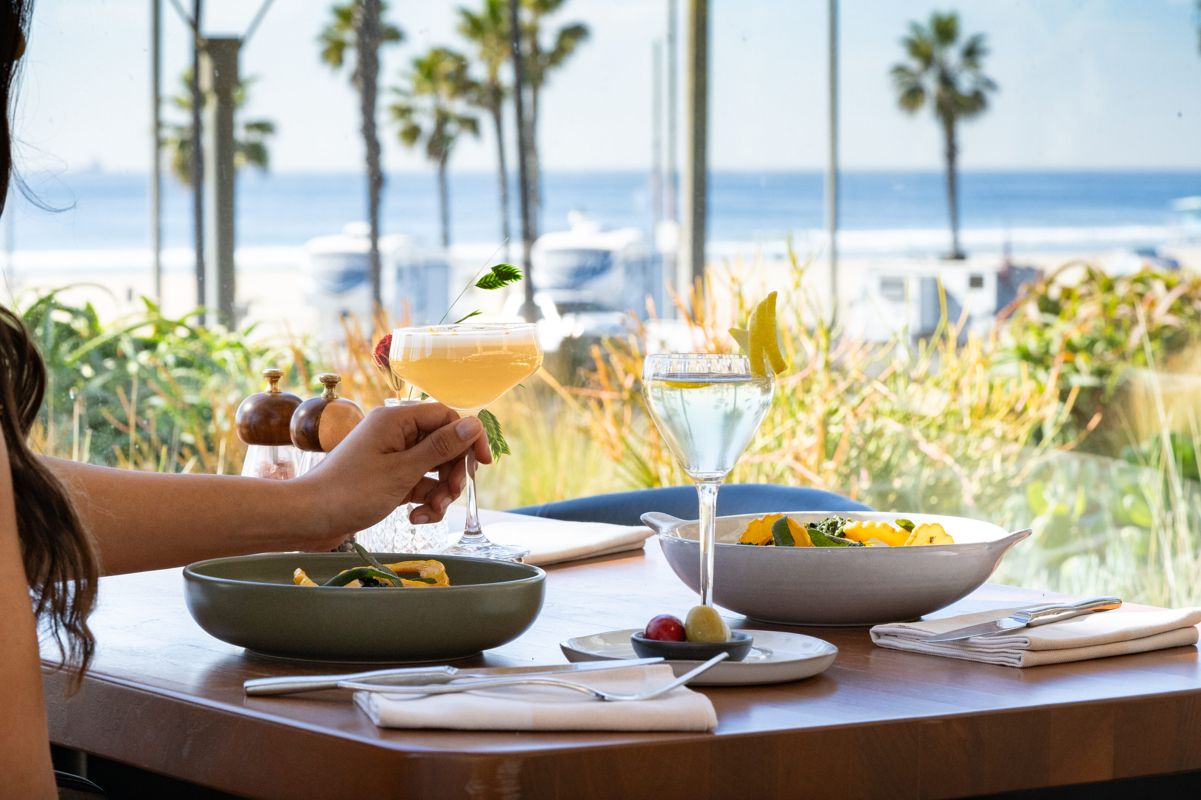 Two people dine at a seaside restaurant table with pasta dishes, cocktails, and a bowl of olives, with palm trees and the ocean visible in the background.