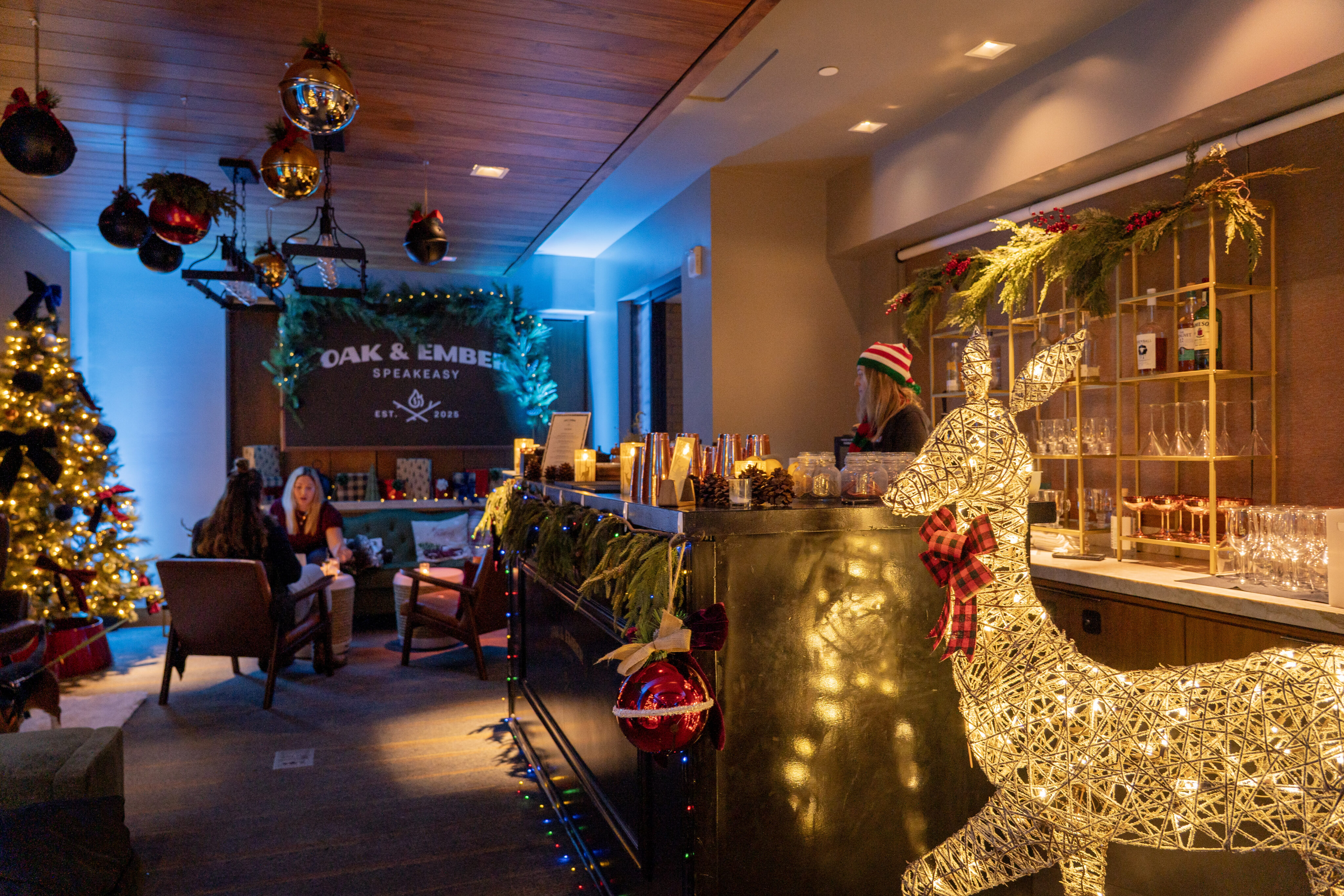 A warmly lit Holiday Speakeasy lounge, adorned for Christmas with a glowing reindeer, festive garlands, ornaments, and a tree; people are seated and talking near an inviting "Oak & Ember" sign.