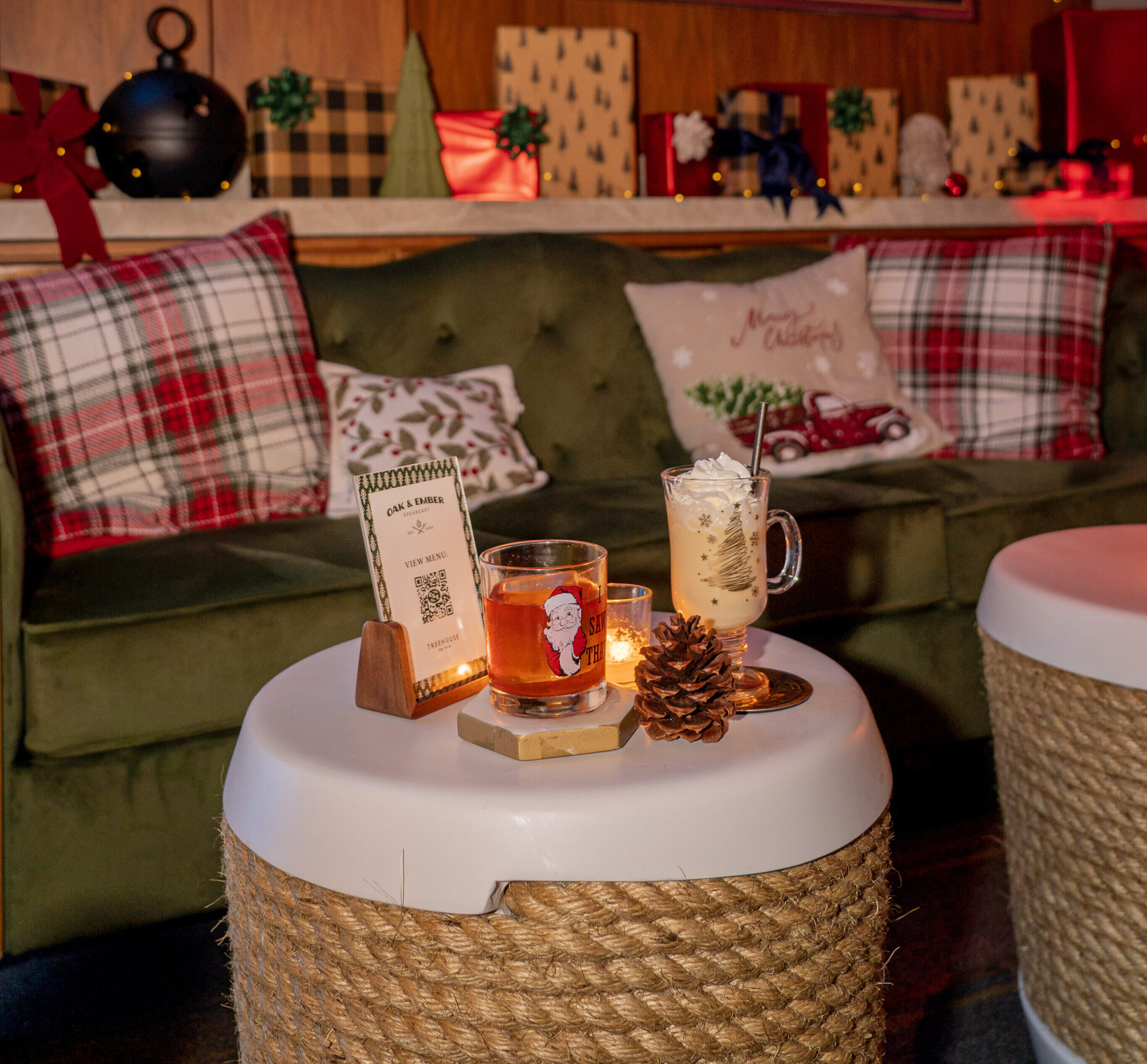 Two festive cocktails on a table in front of a green couch with holiday décor, beneath a sign reading "Oak & Ember Speakeasy Est. 2025," set the perfect scene for a cozy Holiday Speakeasy gathering.