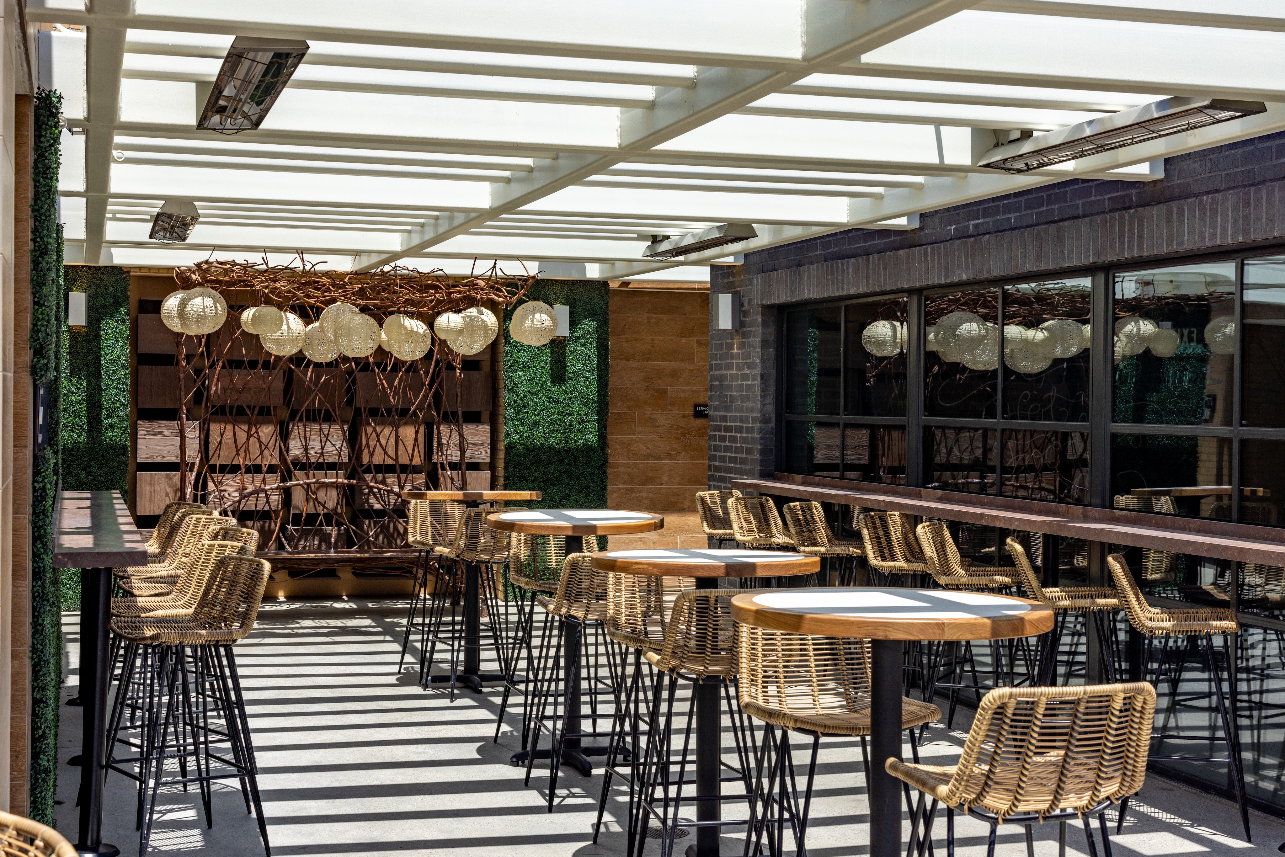 Outdoor patio with wicker chairs and round tables under a white pergola, ideal for celebrations, featuring decorative hanging lights and a mirrored wall reflecting the scene.