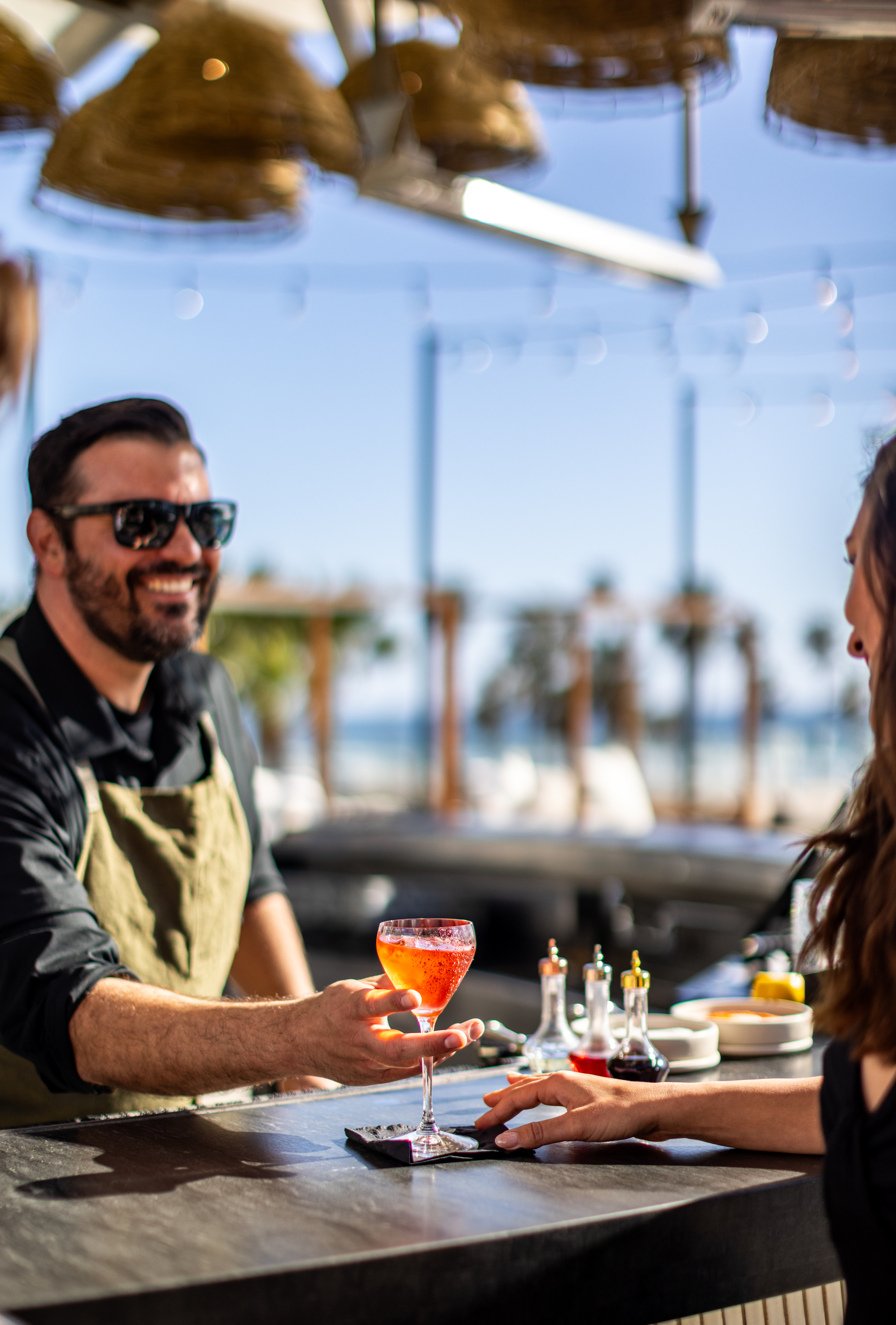 A bartender in sunglasses serves a pink cocktail to a woman at an outdoor bar during Industry Nights, with sunlight and palm trees creating a vibrant, tropical vibe.