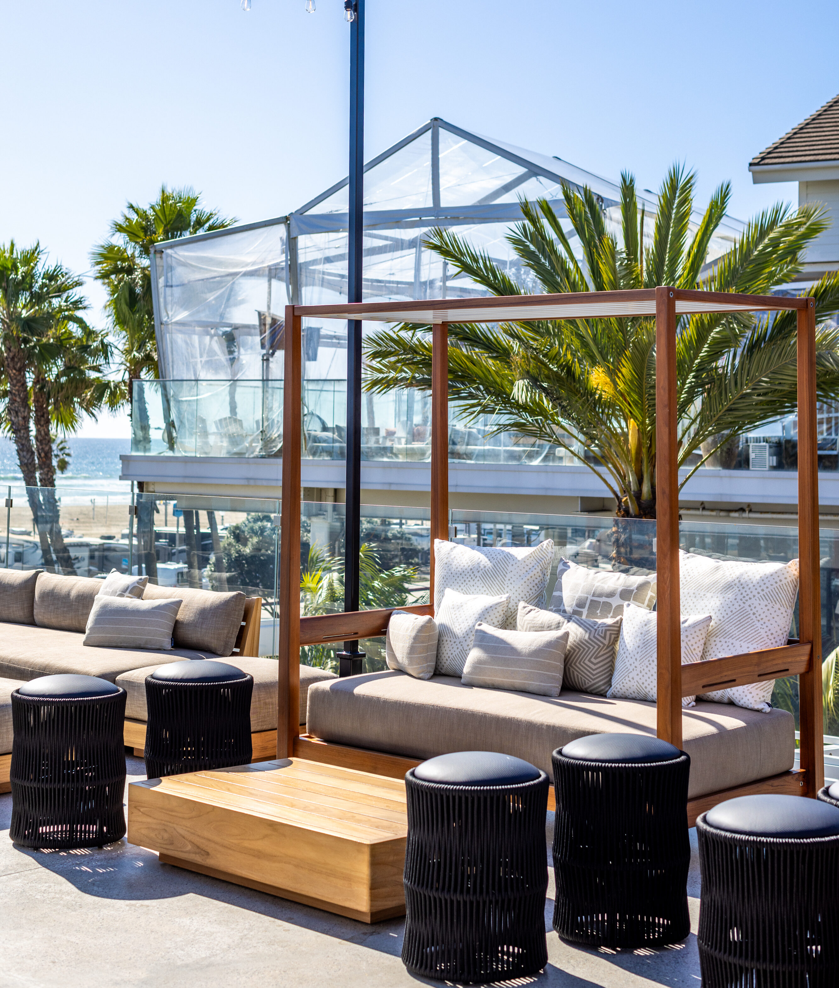 Outdoor lounge area with cushioned sofas, wooden tables, black stools, string lights overhead, palm trees, and a glass structure in the background on a sunny day.