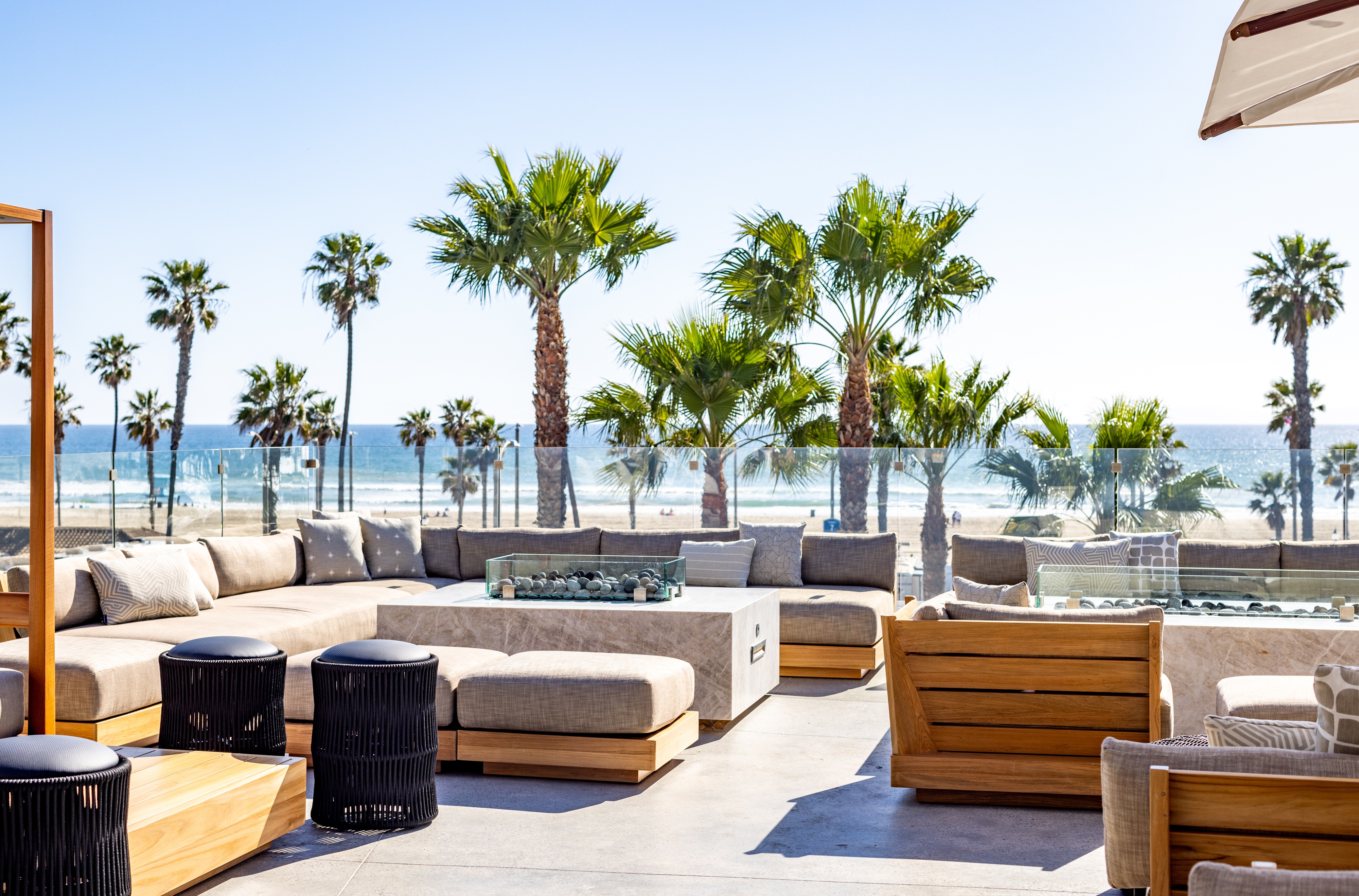 Outdoor lounge area with wooden furniture and cushioned seating overlooks a sandy beach and ocean, with palm trees and clear blue sky in the background.