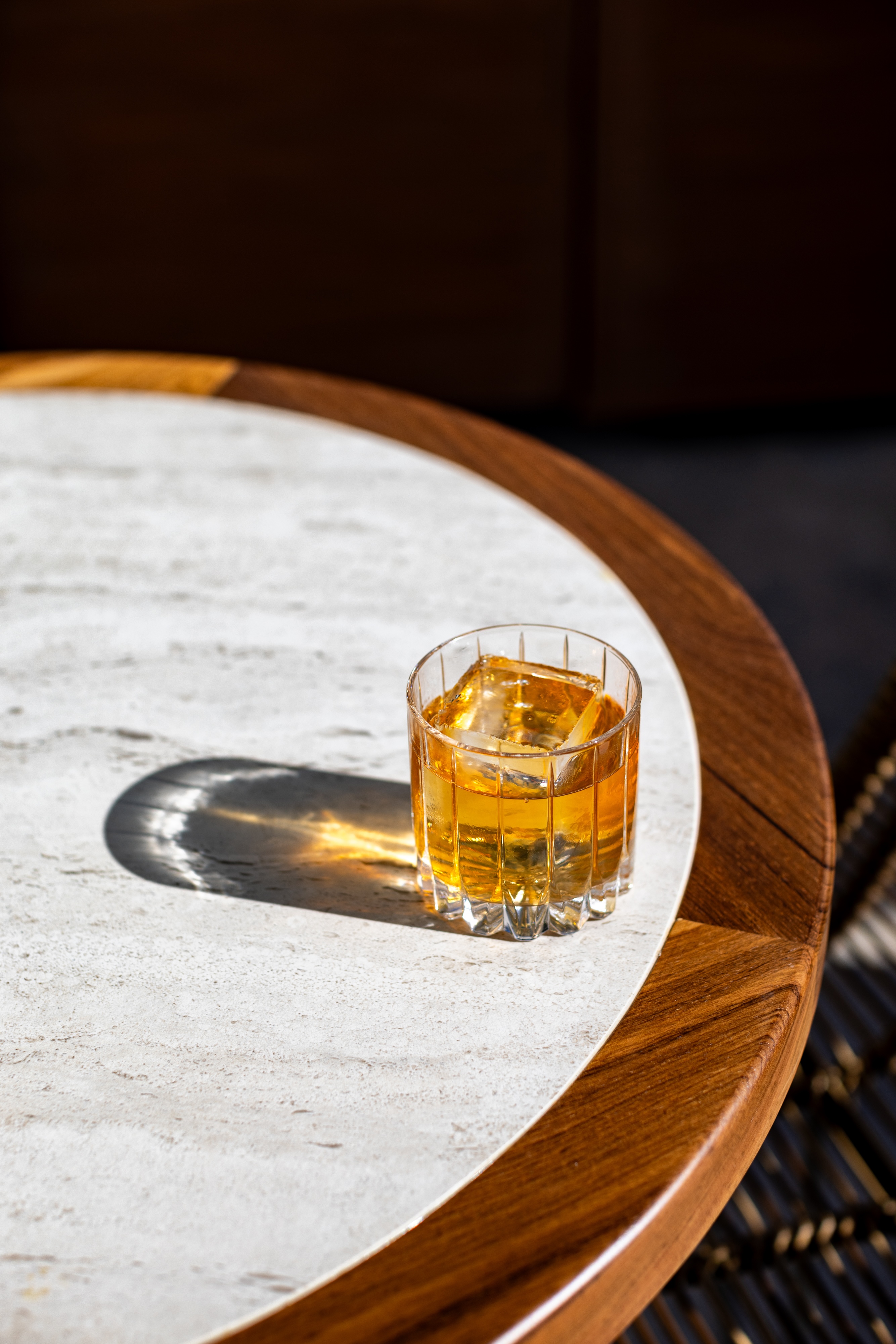 A glass of whiskey with a large ice cube sits on a round marble and wood table, casting a shadow in bright sunlight.