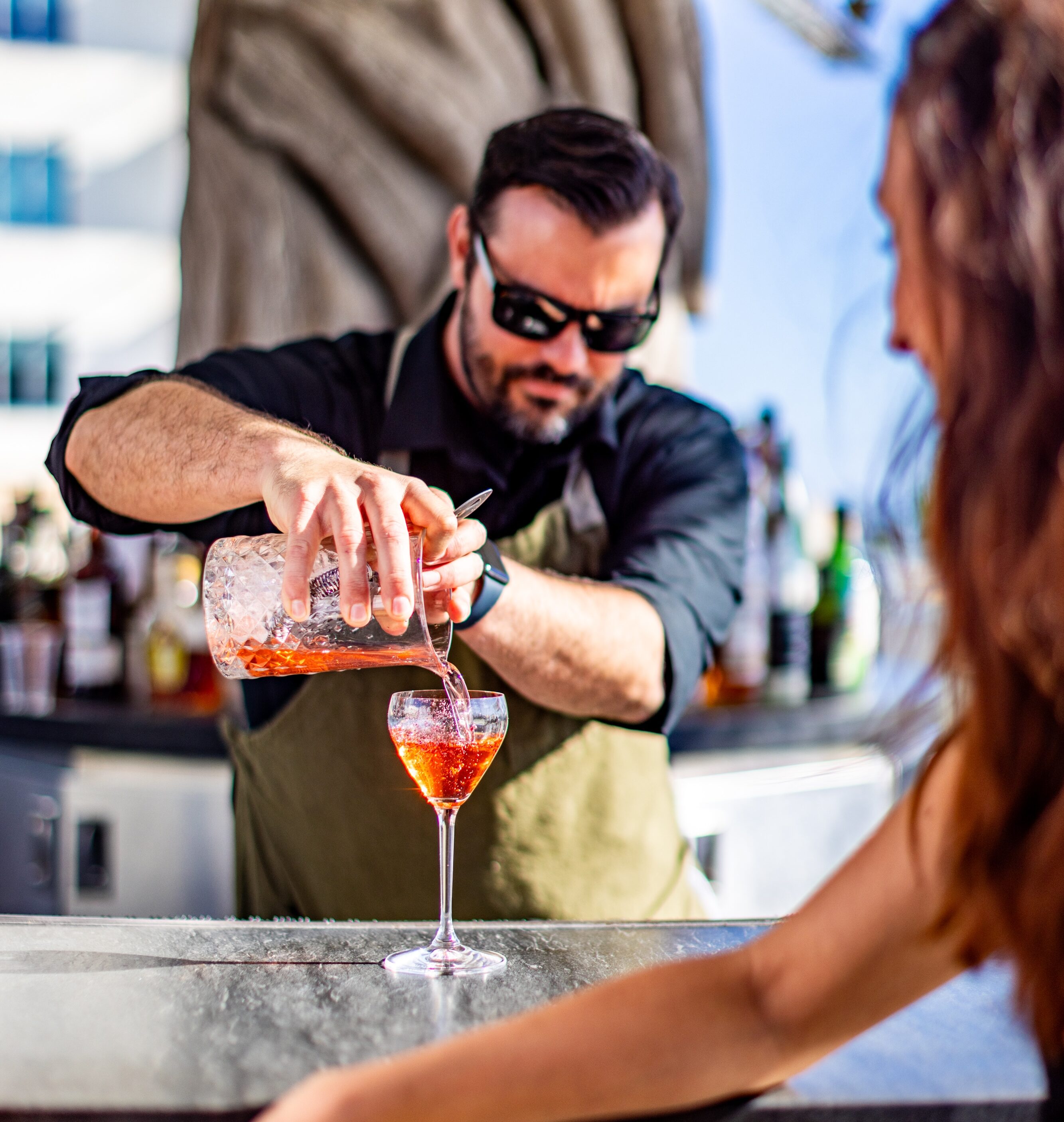 Bartender wearing sunglasses pours a red cocktail into a glass at an outdoor bar while a woman with long hair leans on the counter.