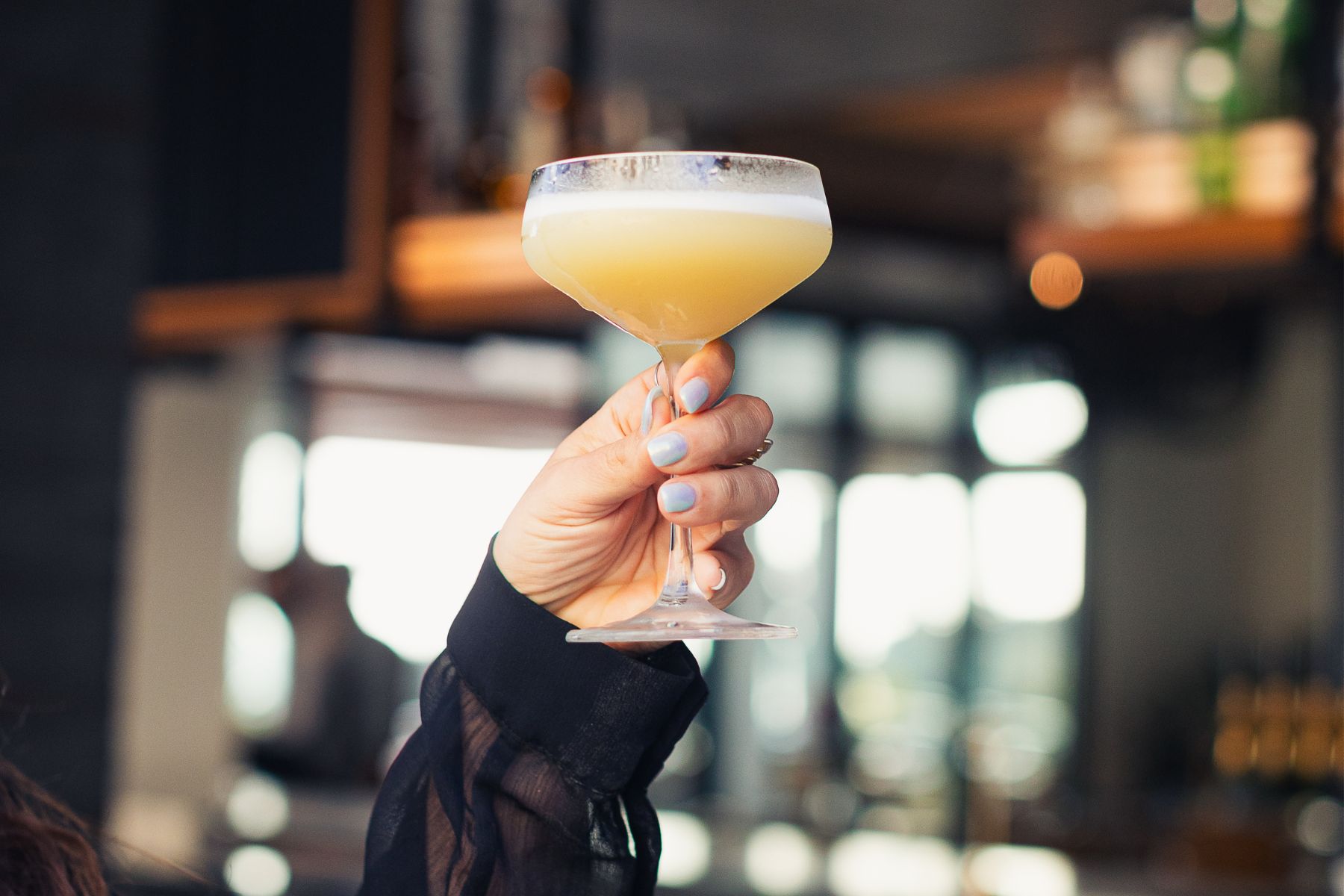 A hand with light blue nail polish holds up a coupe glass filled with a yellow cocktail in a bar setting.