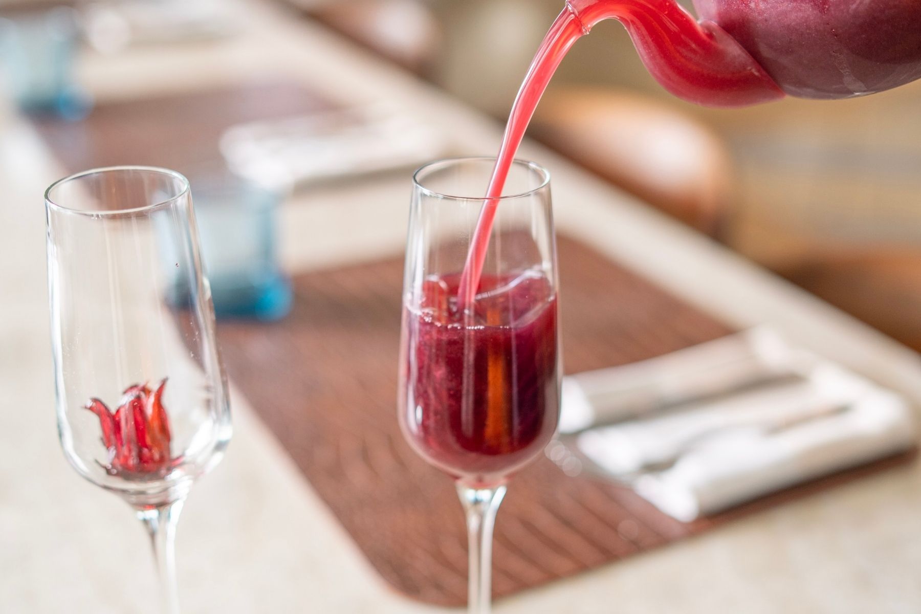 A close-up of a Love Potion-inspired red beverage being poured into a champagne flute, with another empty flute containing a red flower, on a set dining table.