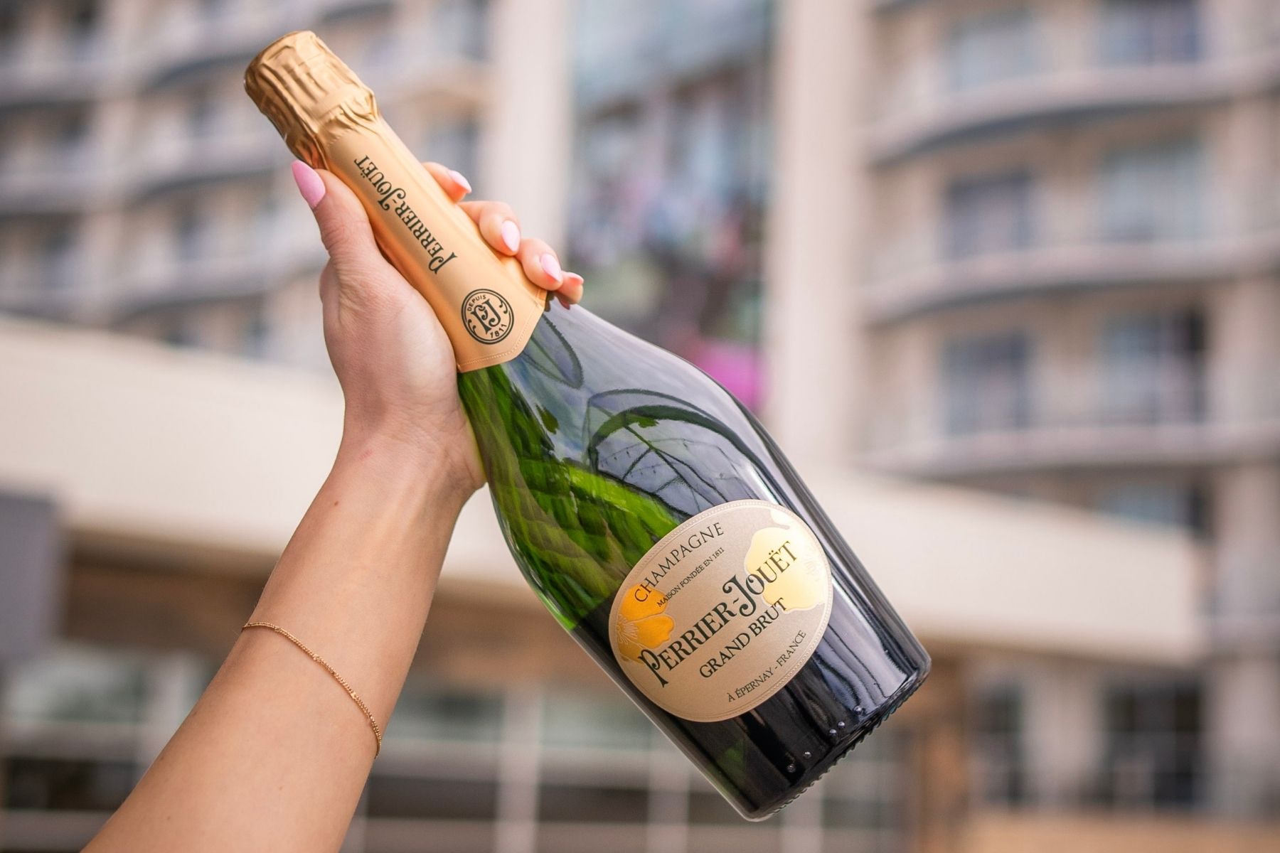 A hand holds a bottle of PJ's champagne outdoors, perfect for a Galentine's Brunch, with blurred modern building balconies in the background.
