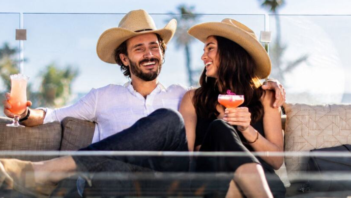 A man and woman wearing cowboy hats sit on an outdoor couch, smiling and holding cocktails, enjoying a festive Codigo Country Night with palm trees visible in the background.