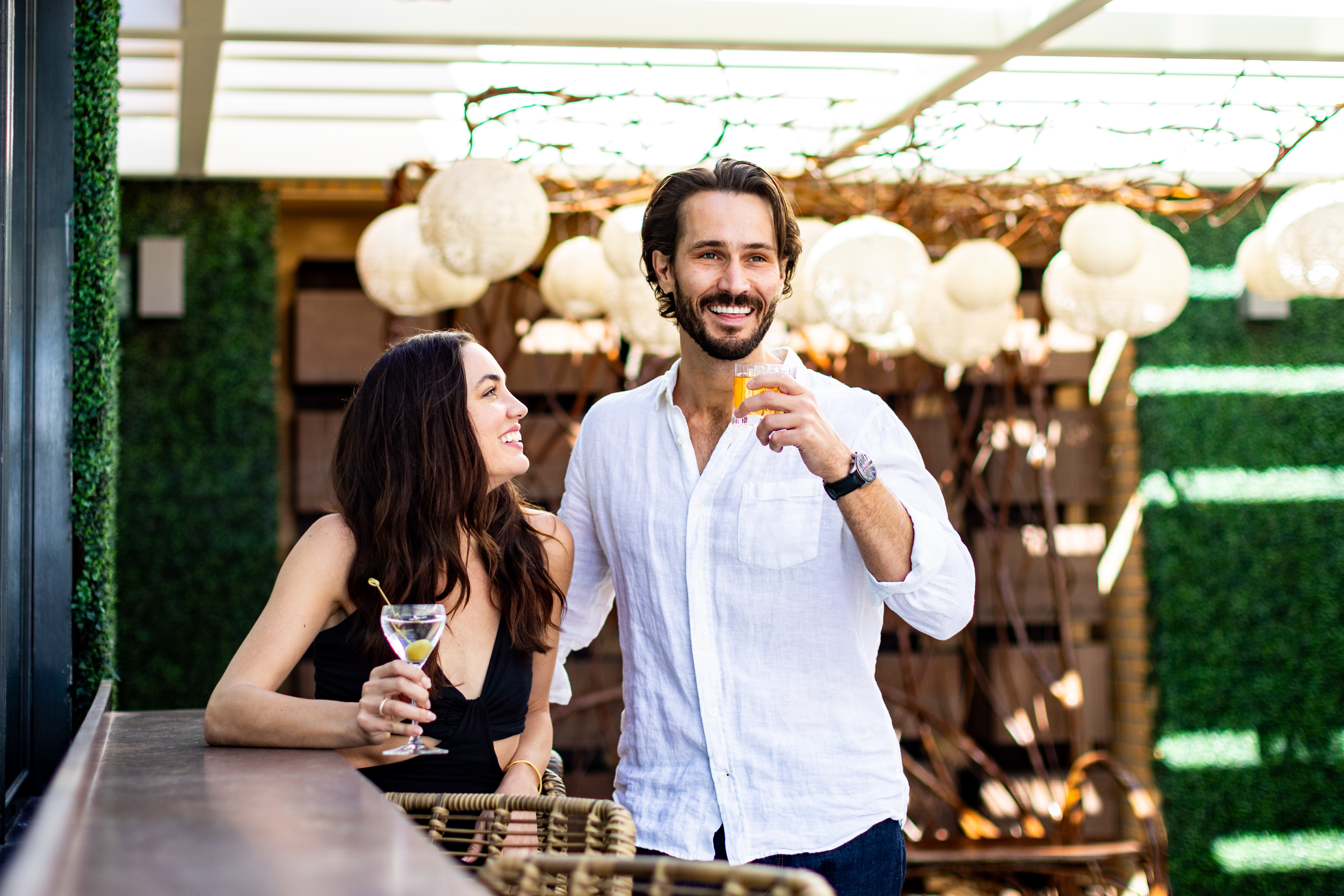A man and woman stand at an outdoor bar, smiling and holding drinks, with hanging lights and greenery in the background.
