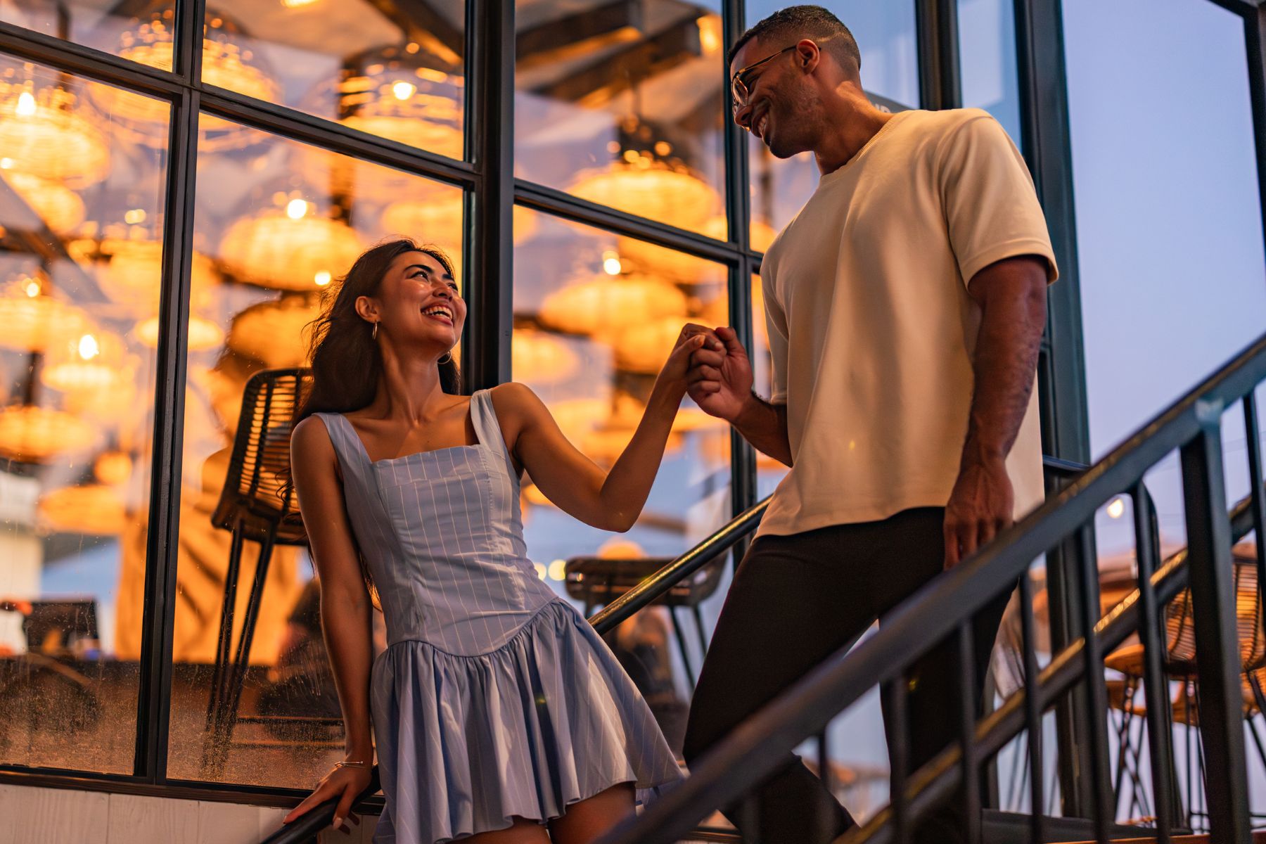 A woman in a light dress and a man in a light shirt hold hands and smile at each other on stairs, with warm hanging lights in the background, capturing the magic of a late night happy hour together.