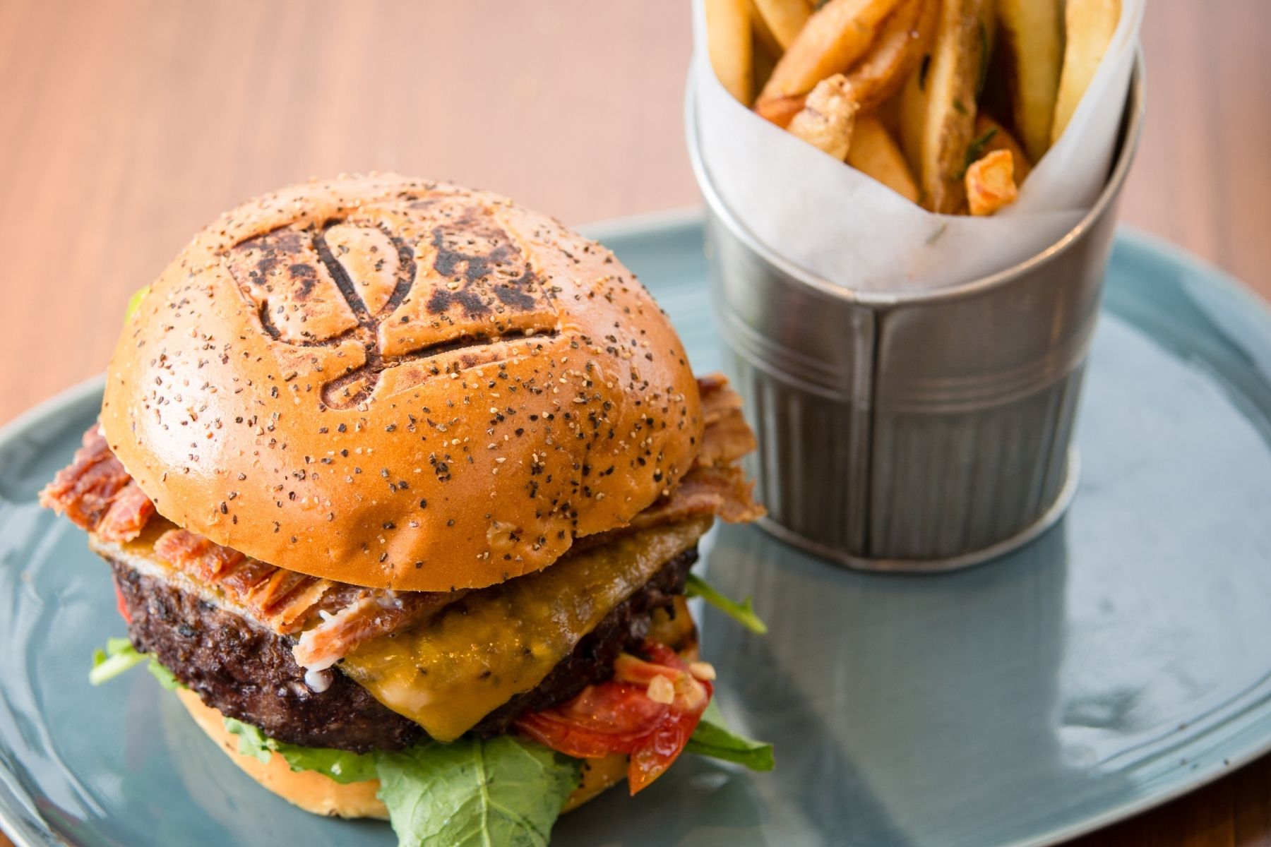 Savor a Burger Week favorite: a cheeseburger with bacon, lettuce, and tomato on a poppy seed bun, served with a side of French fries in a metal container on a blue plate—an Orange County classic.
