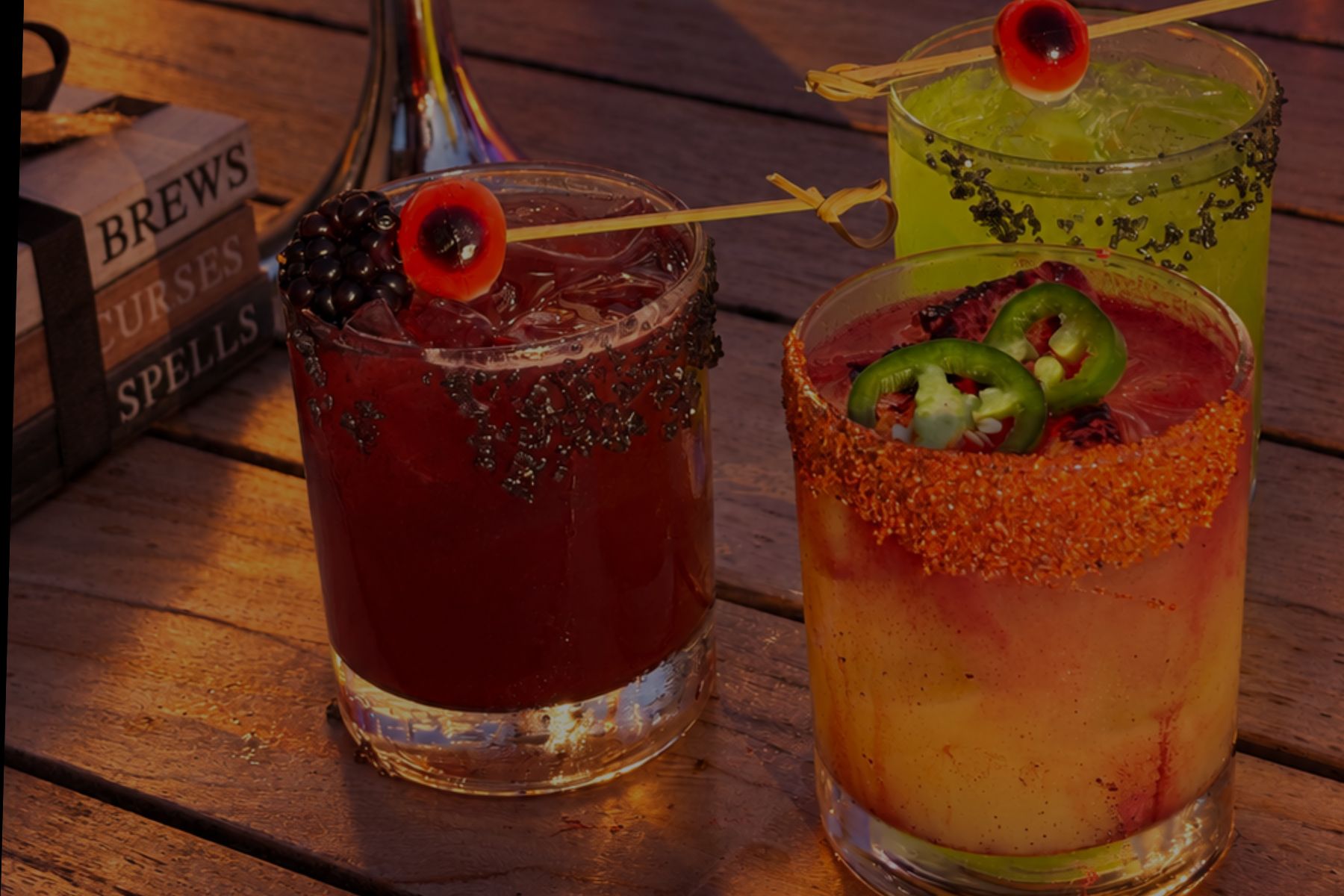 Three cocktails in glass tumblers on a wooden table, featuring fruit and chili garnishes.
